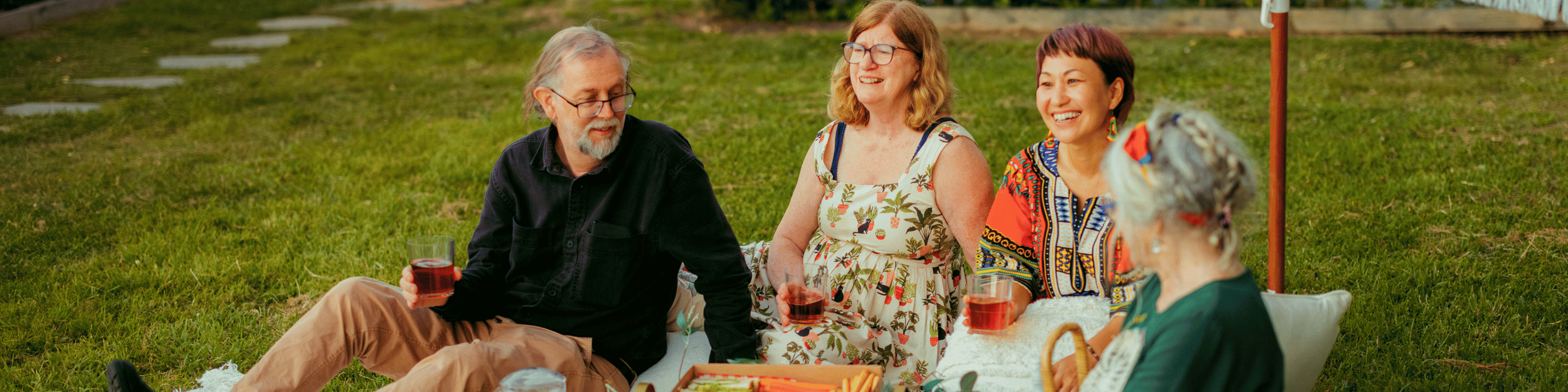 Four adults sitting on a blanket on the grass at a picnic, smiling and holding glasses of red wine, with a woman knitting.