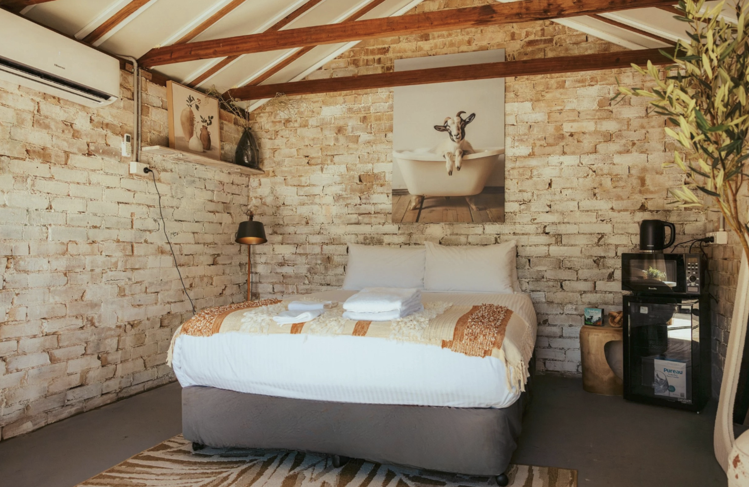 Cozy bedroom with a brick wall, bed with white linens and orange blanket, wall art of a goat in a bathtub, plants, and various small appliances.