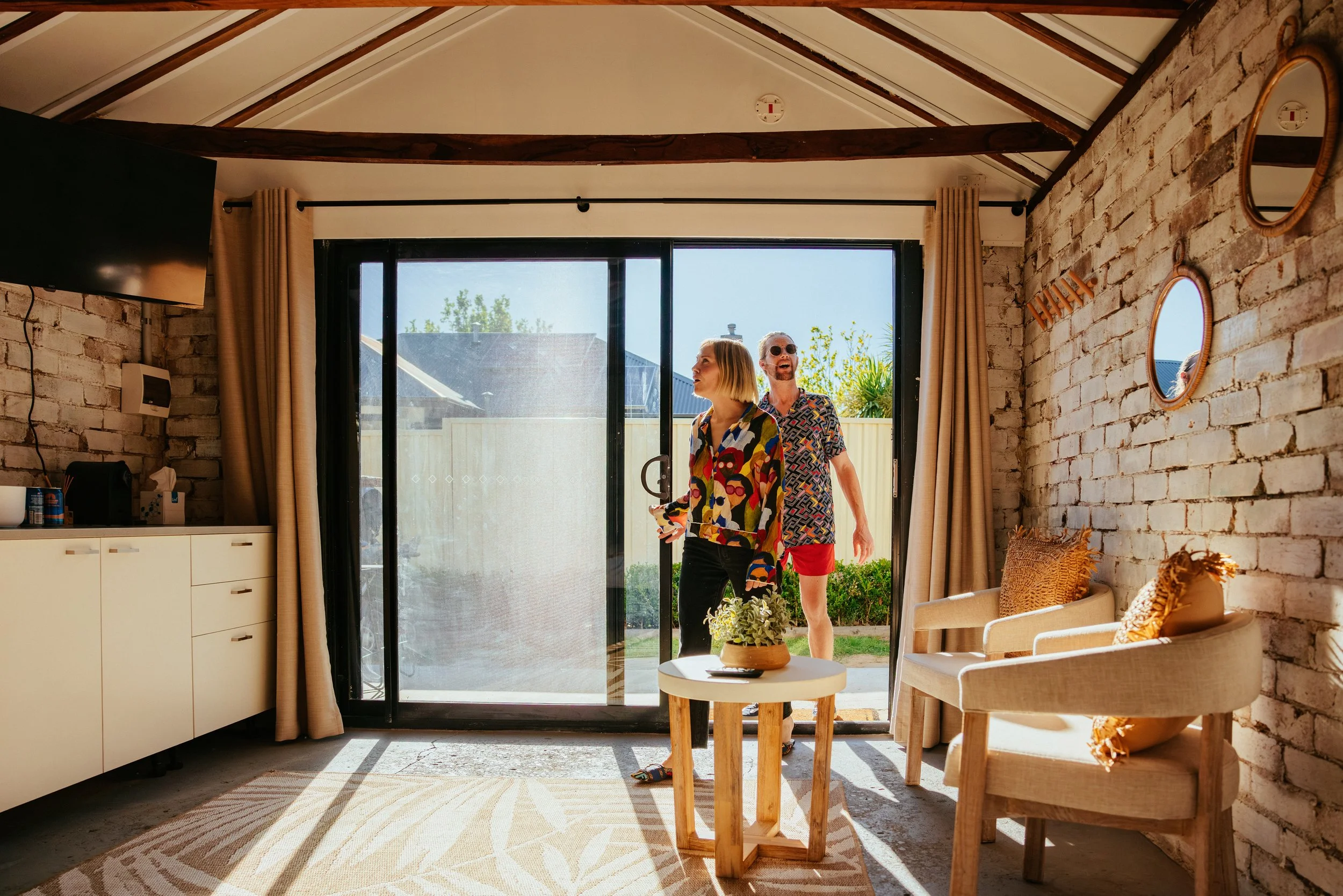 A modern living room with brick walls, beige curtains, and a sliding glass door leading to the outdoor yard. Two people, a woman and a man, are standing near the door, looking outside and smiling.