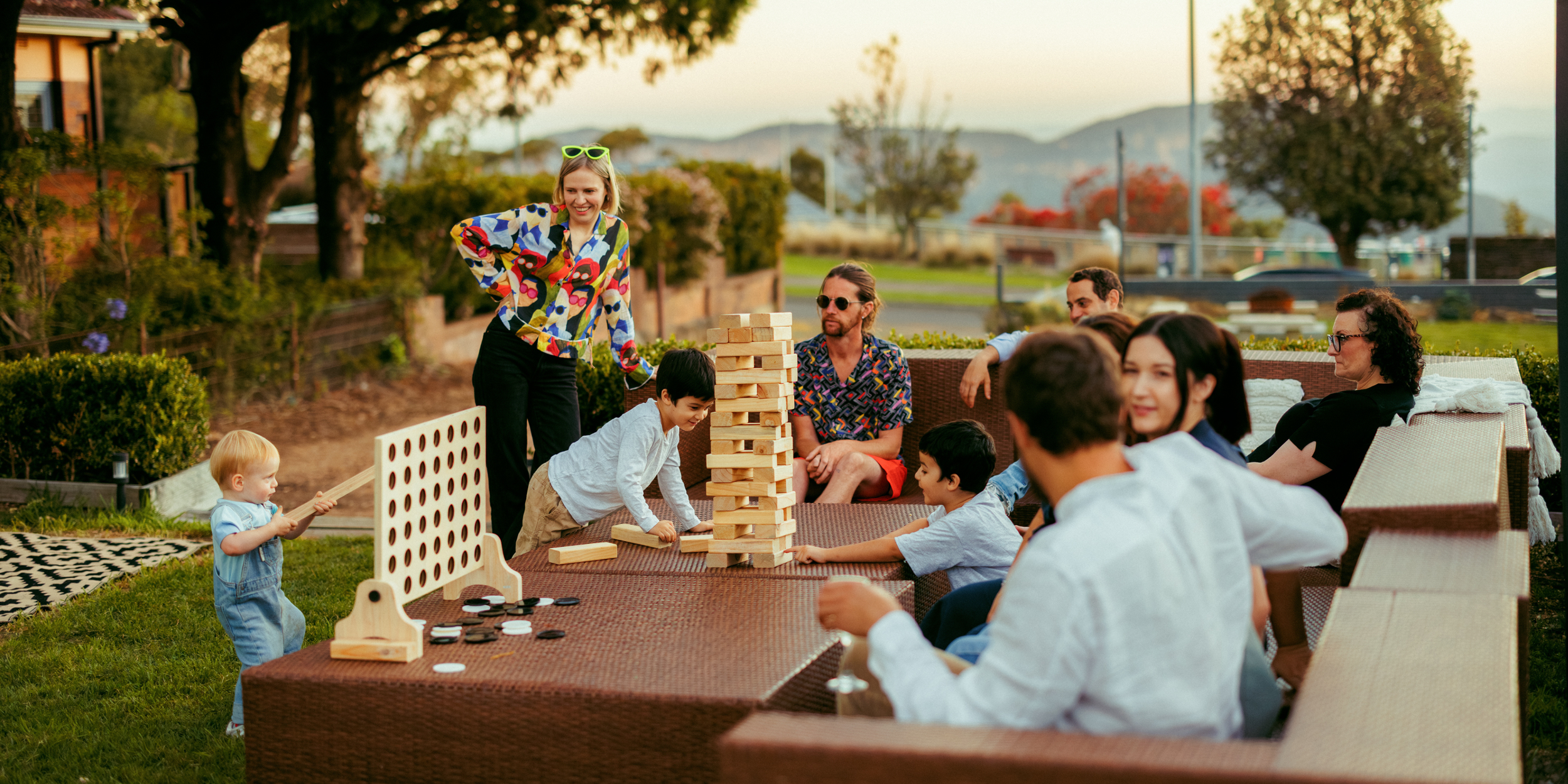 A group of people, including children and adults, playing a giant outdoor Jenga game at a backyard gathering during sunset with trees and a mountain in the background.