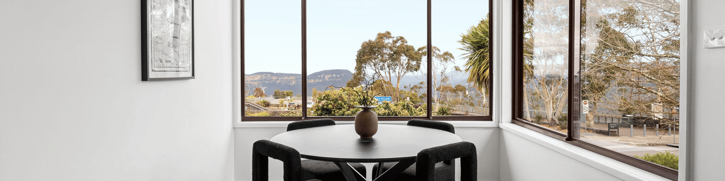 A round dining table with a vase of branches, surrounded by four black chairs, is in a corner with large windows showing trees and mountains outside.