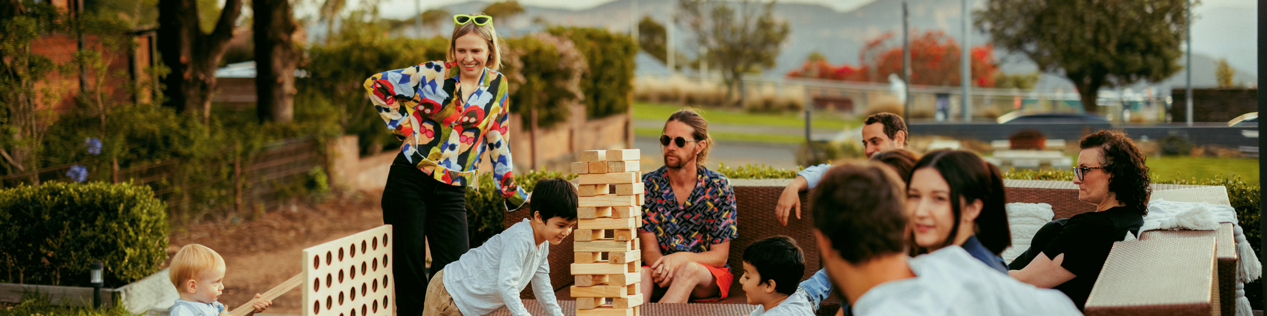 A group of people playing oversized Jenga outside in a backyard. A woman in a colorful blouse is about to remove a block, while children and adults watch and laugh.