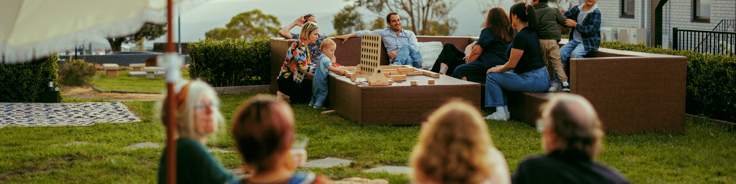 Group of people, including children and adults, sitting and socializing outdoors on a grassy area with patio furniture and trees in the background.