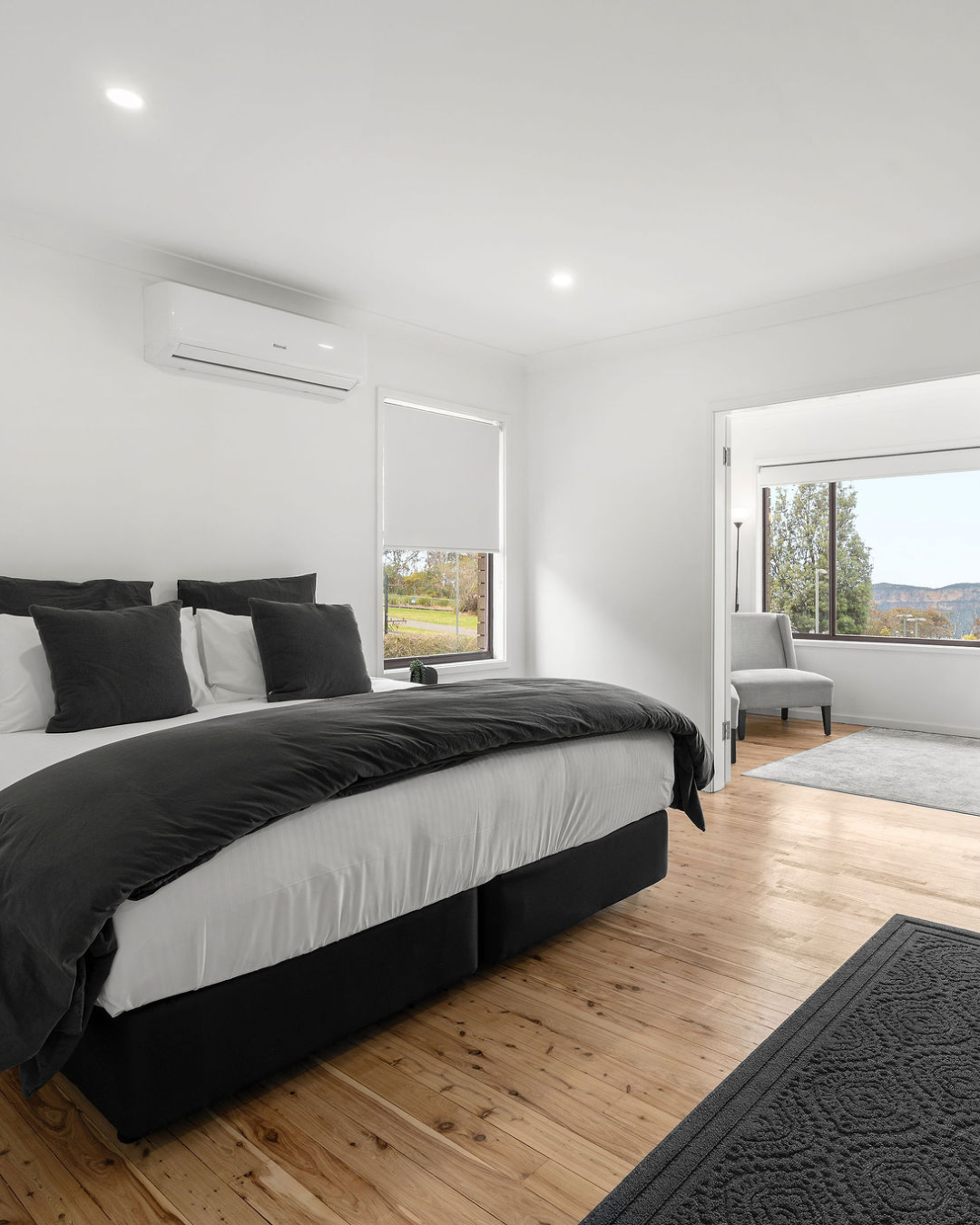 A modern bedroom with white walls, a black and white bed, large windows showing a scenic outdoor view, and a gray armchair by the window.