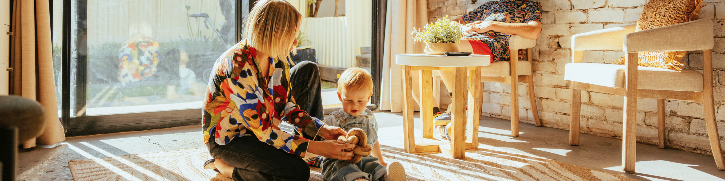 A woman and a young boy sitting on a beige rug near a sliding glass door, playing with a toy shell. Sunlight streams into the room, which has a brick wall, wooden furniture, and a potted plant on a round table.