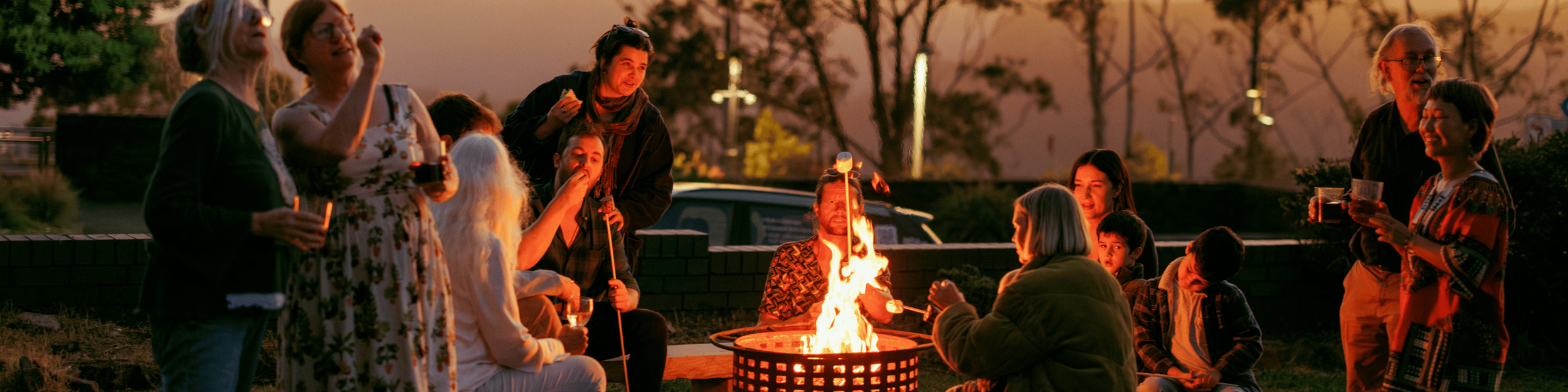 Group of people gathered around a campfire outdoors during sunset, enjoying marshmallows and drinks.