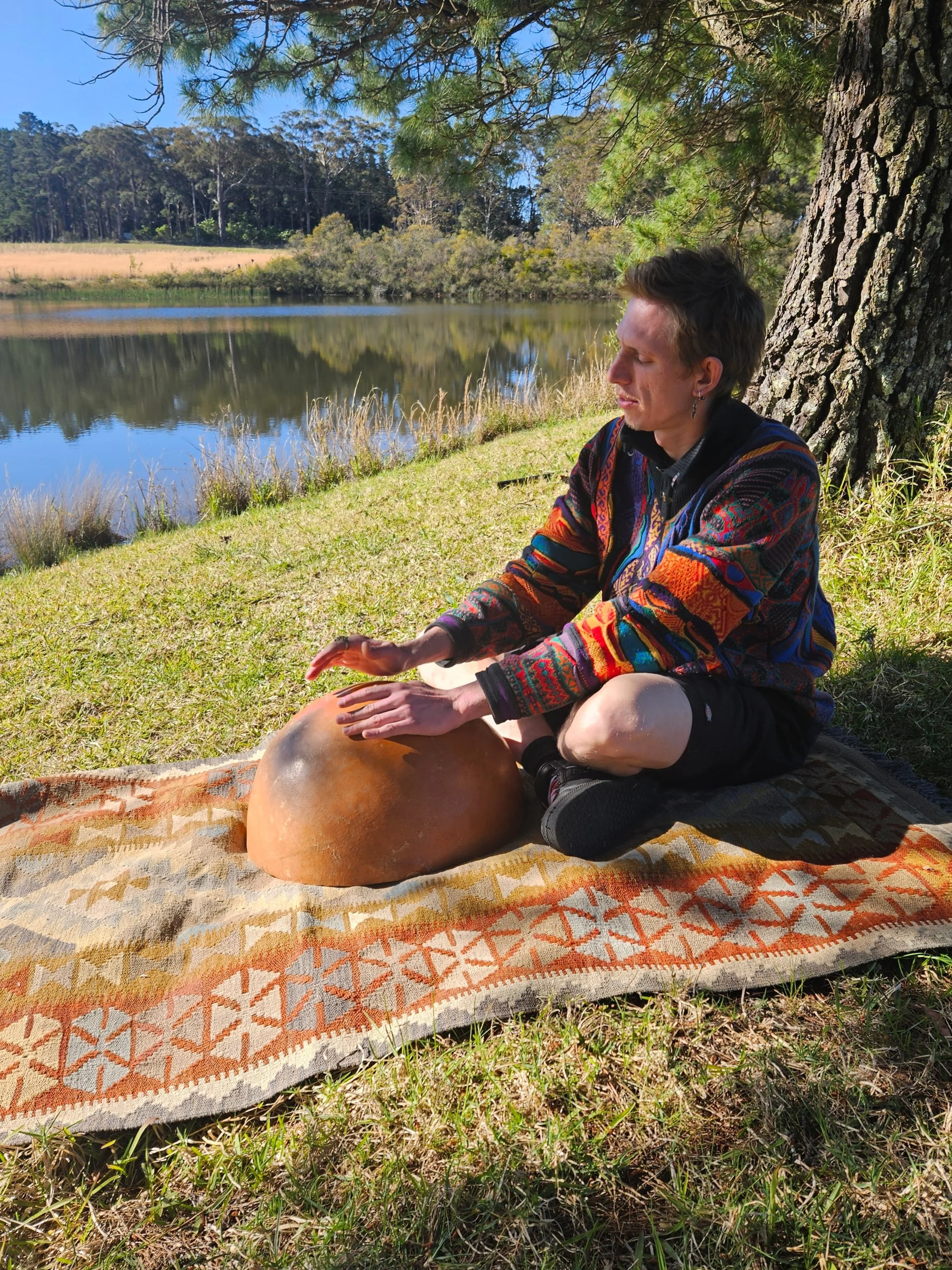 Person sitting on a colorful rug outdoors, practicing meditation by a lake shaded by trees.