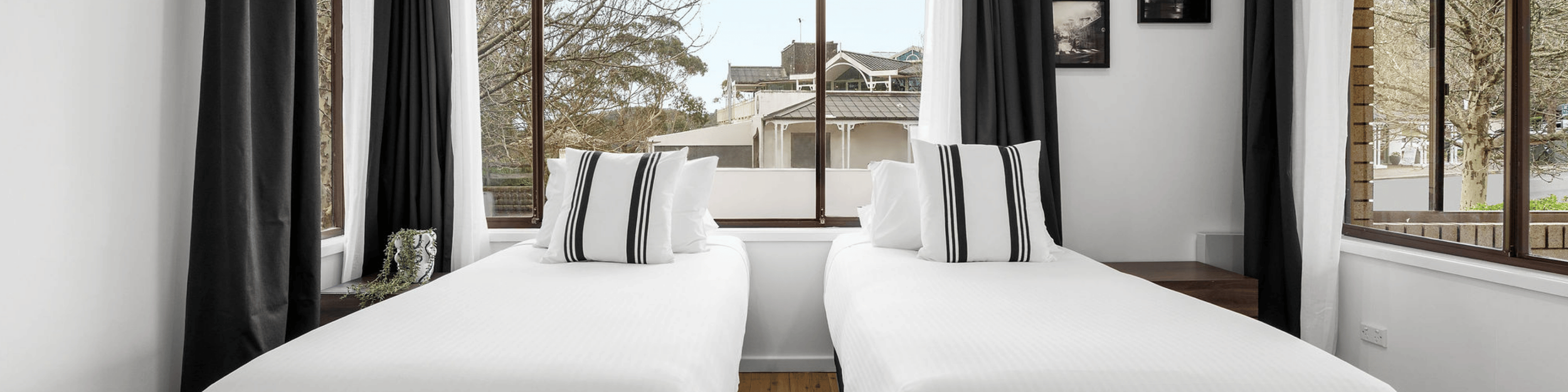 Room with two twin beds, each with white bedding and black-and-white striped pillows, against large windows with black and white curtains, overlooking neighboring houses and trees.
