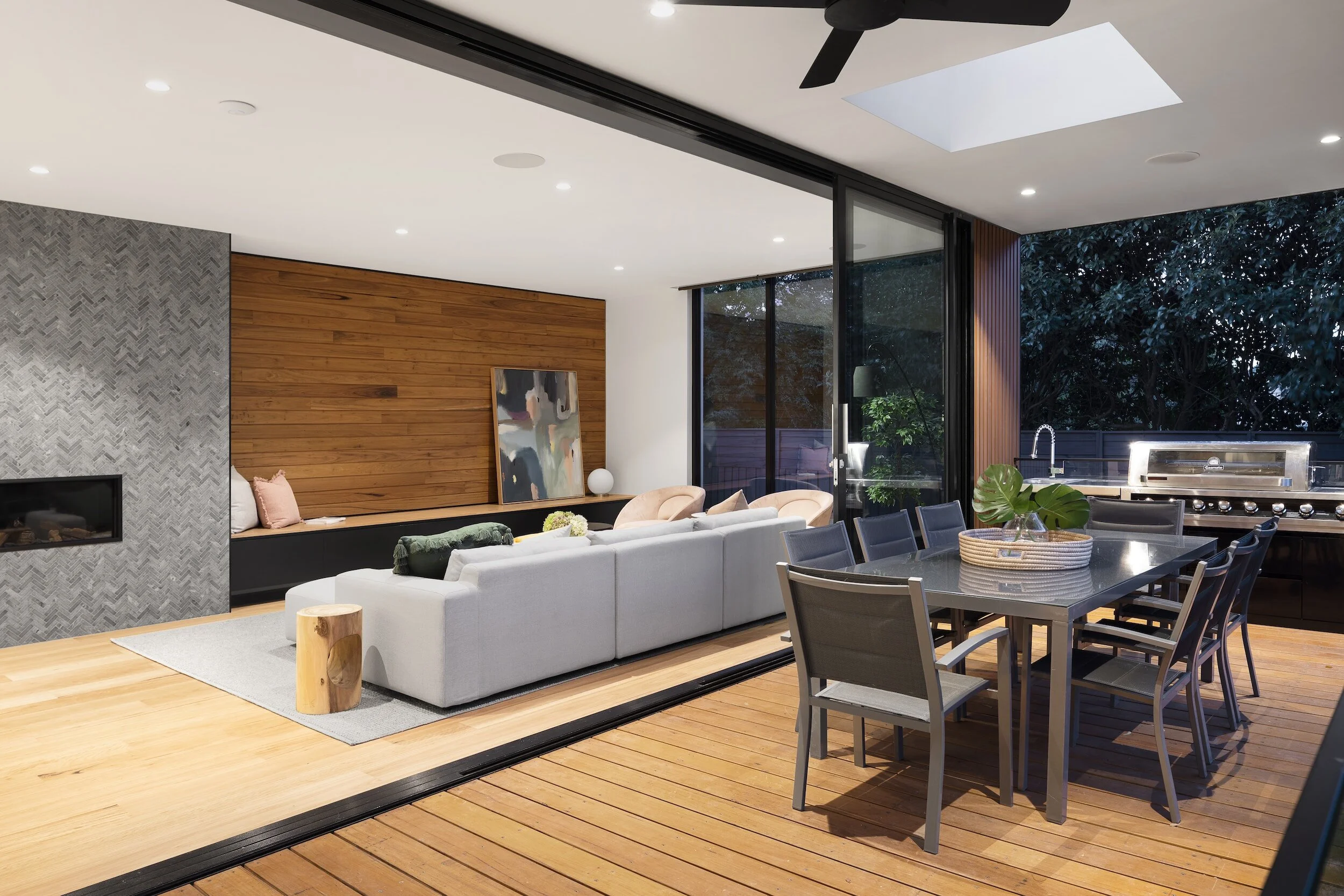 Modern open-concept living and dining area with a gray sectional sofa, wood panel wall, contemporary painting, outdoor dining table, grill, and a large sliding glass door opening to a patio with trees.