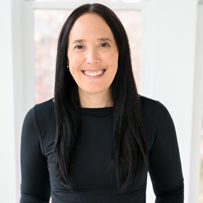 Portrait of a woman with long dark hair smiling, wearing a black top, in front of a window with a blurred background.