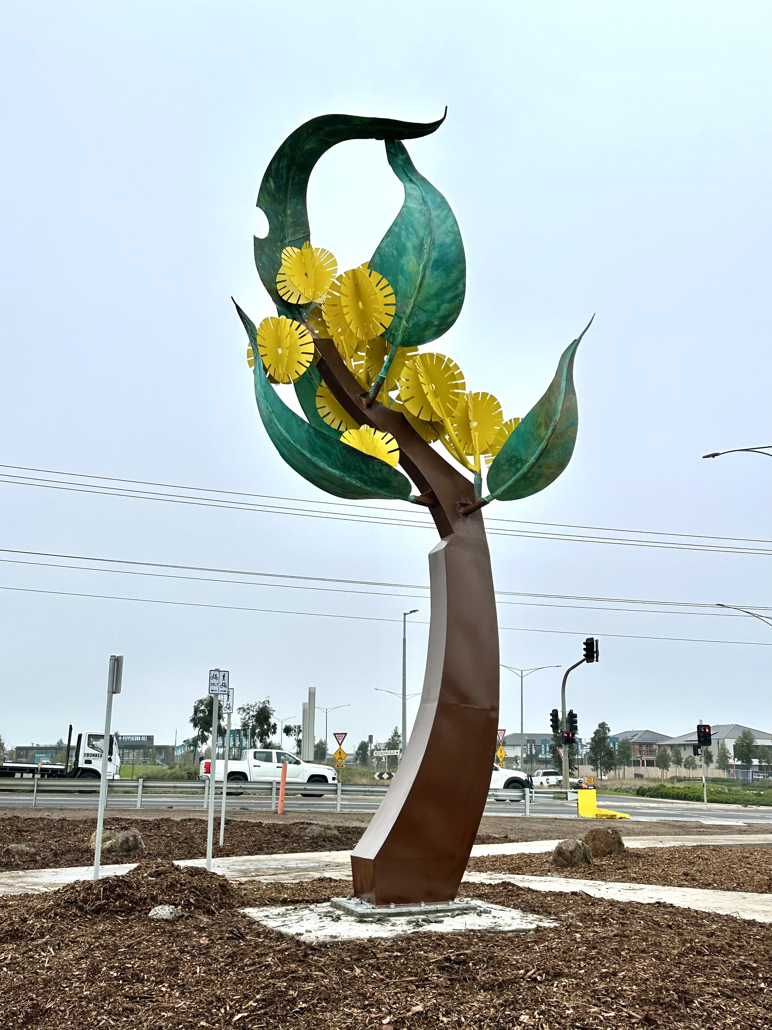 Matilda public sculpture by artist blacksmith David Wood installed at Donnybrook, with golden wattle-inspired steel stem, green leaves and yellow blossoms.