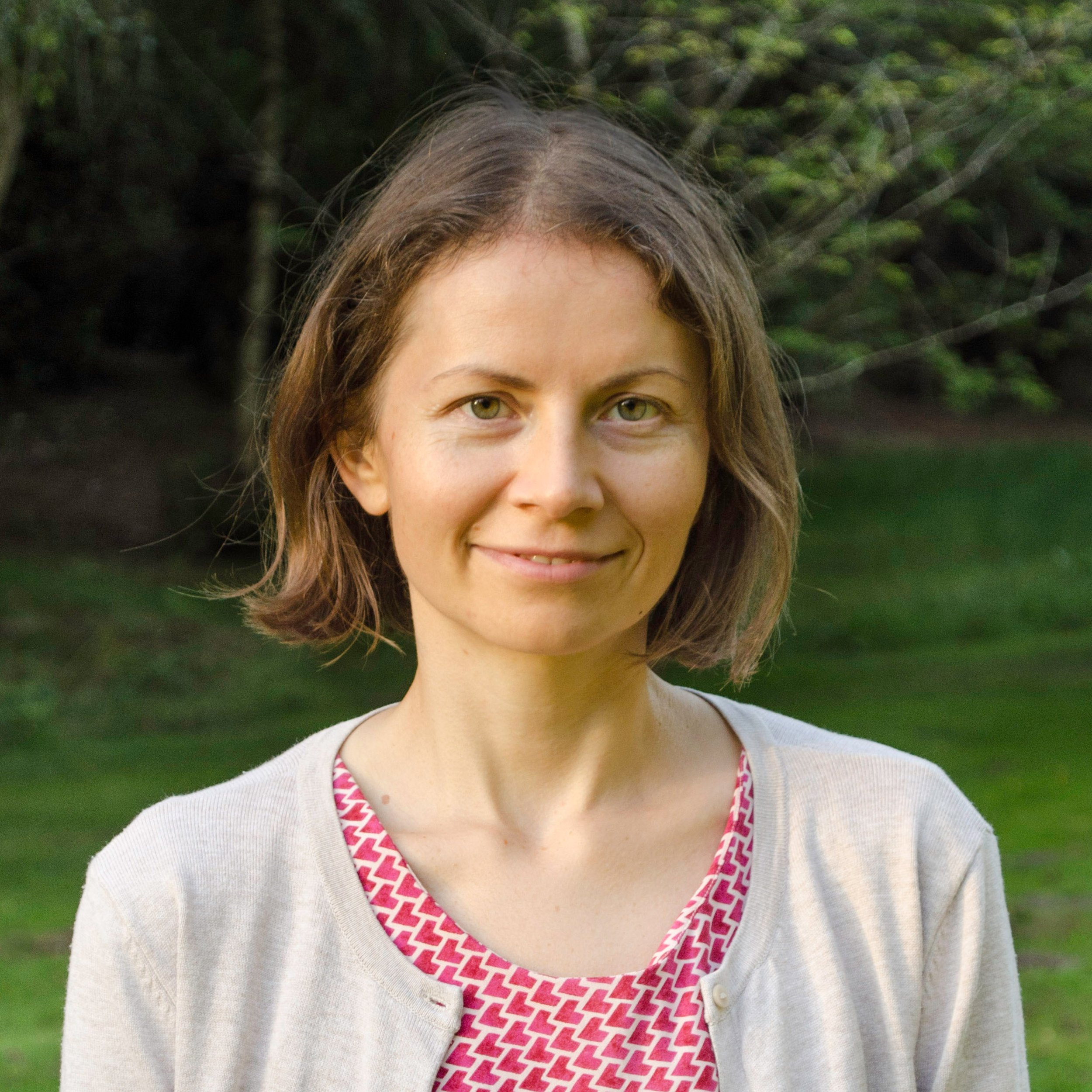 A woman with short brown hair and green eyes smiling outdoors with trees and grass in the background.