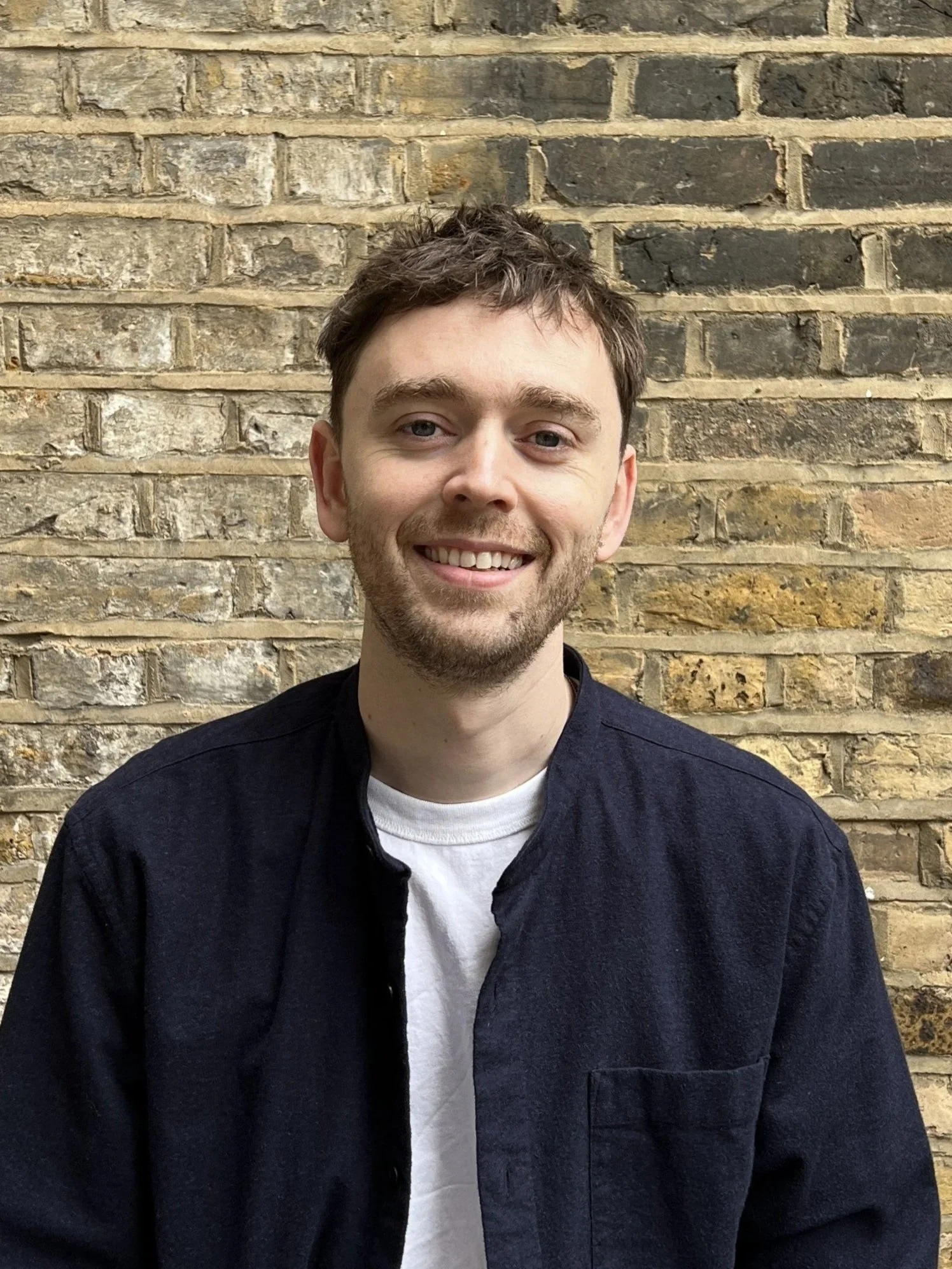 A young man with light brown hair, a beard, and blue eyes smiling while sitting in front of a brick wall.