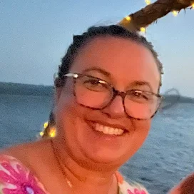 A person smiling and wearing sunglasses taking a selfie at the beach, with the ocean and waves in the background and another person standing near the shoreline.