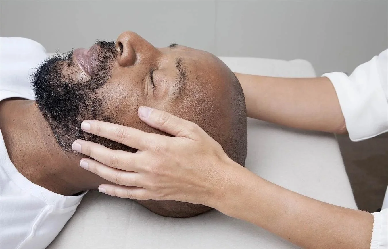 A person receiving a facial massage or treatment while lying down on a treatment bed, with eyes closed and head supported by a therapist's hands.