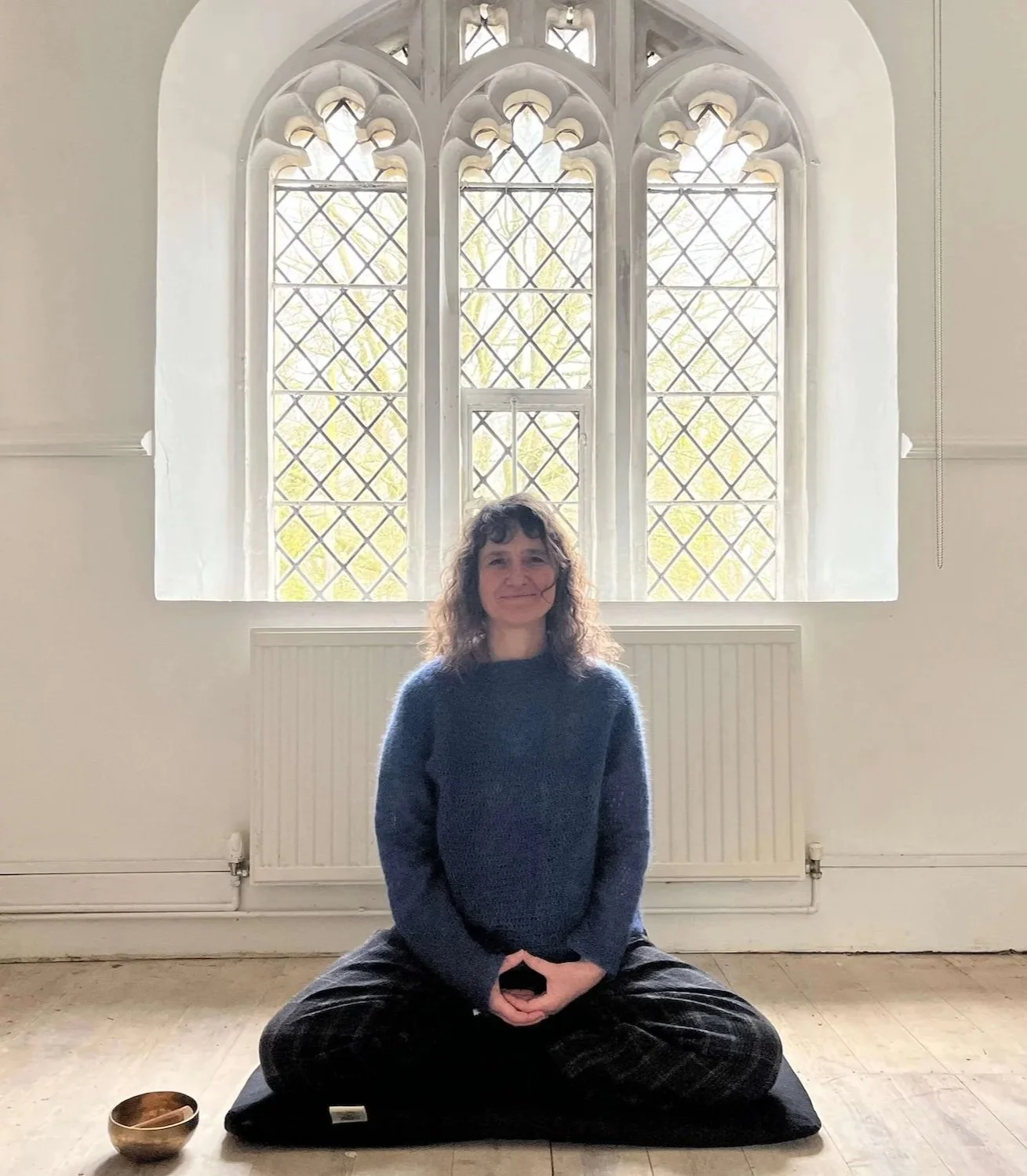 A woman sitting cross-legged on a cushion in a room with large gothic-style windows, wearing a blue sweater and dark pants, with a singing bowl and a small object nearby.