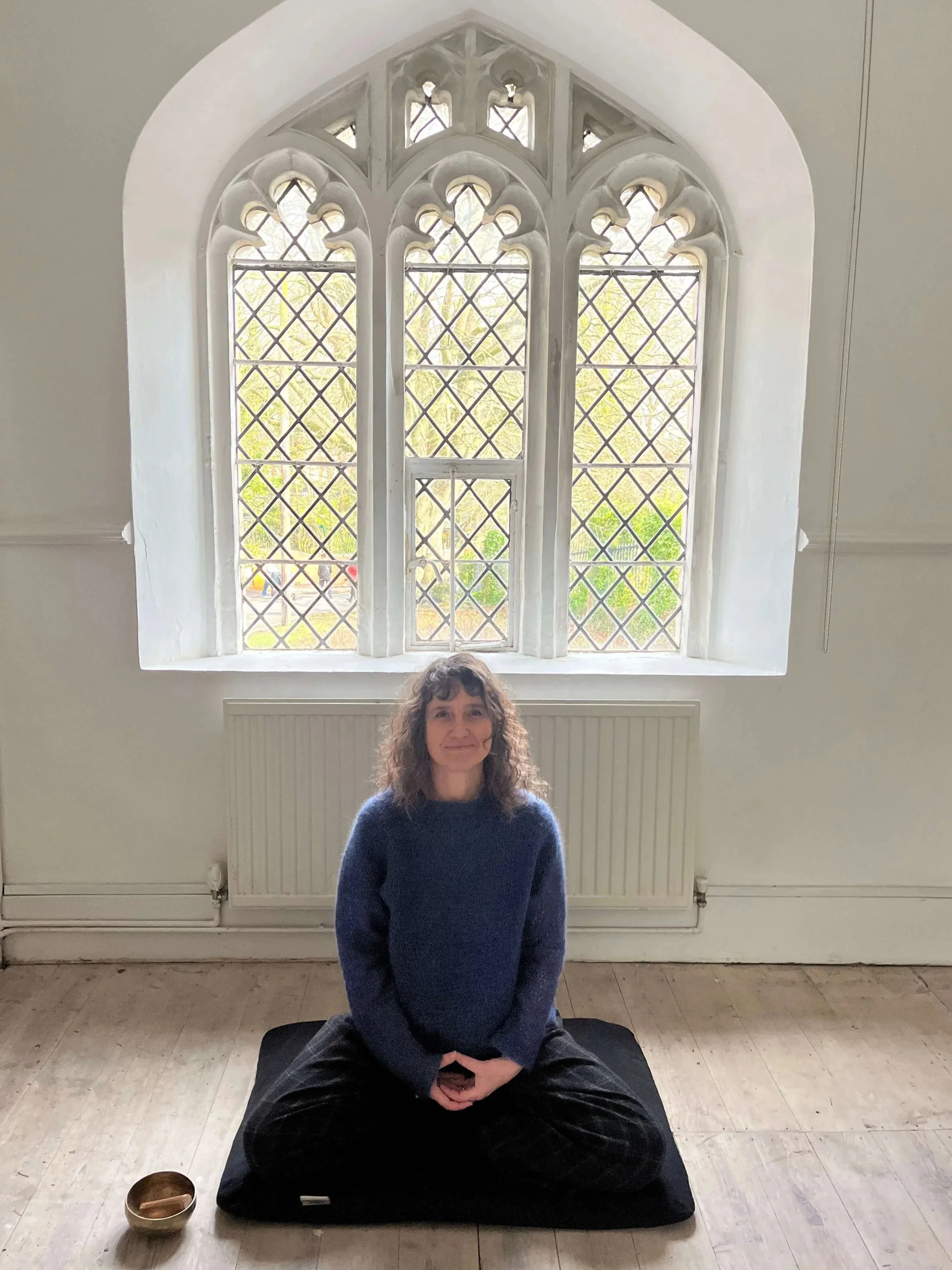 A woman with curly hair sitting cross-legged on a black cushion in a room with a large Gothic-style window behind her, with greenery visible outside. There is a small bowl on the floor to her left.