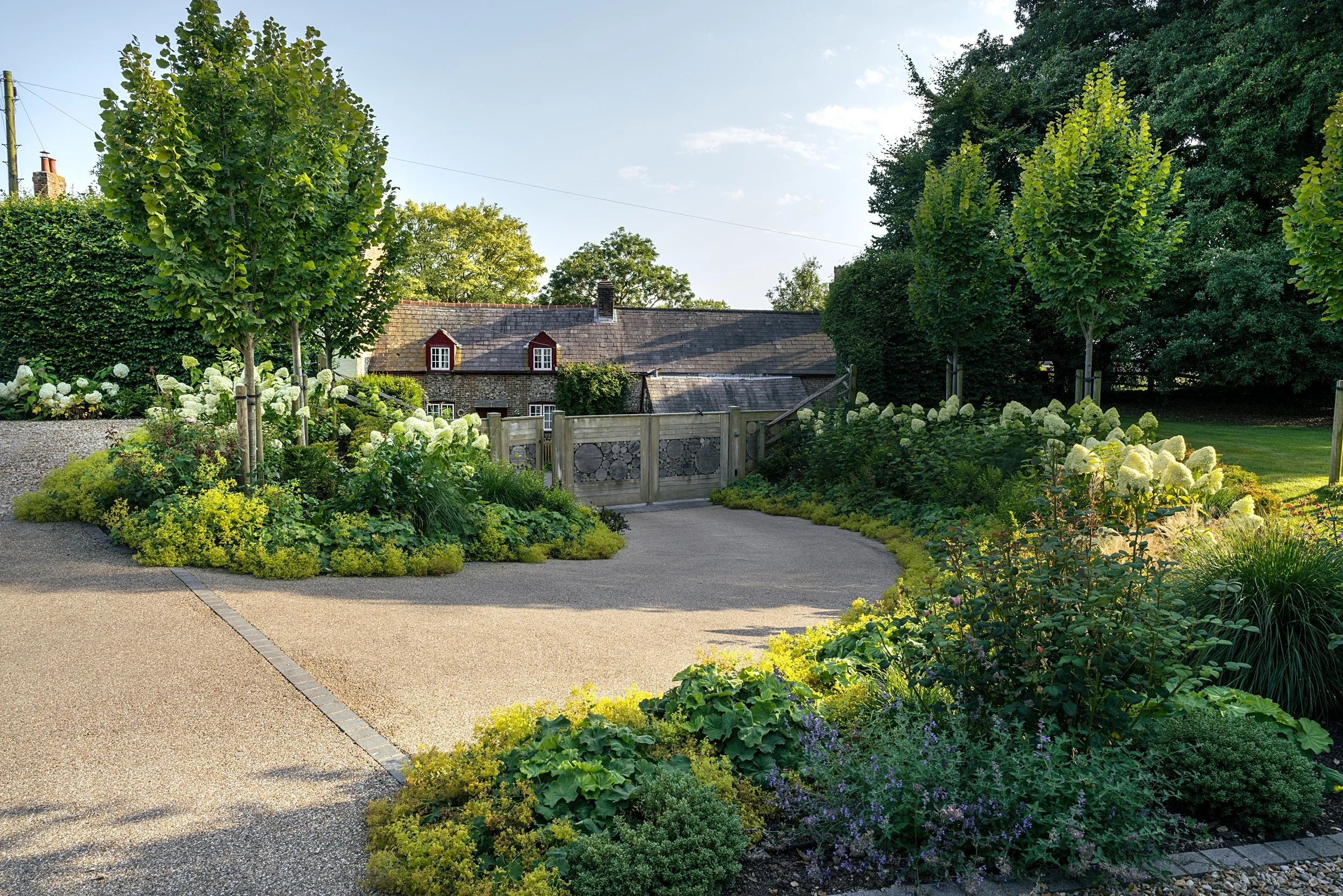 A garden with lush green trees, white flowering bushes, and various plants along a paved walkway and stone wall, with a house roof and sky in the background.