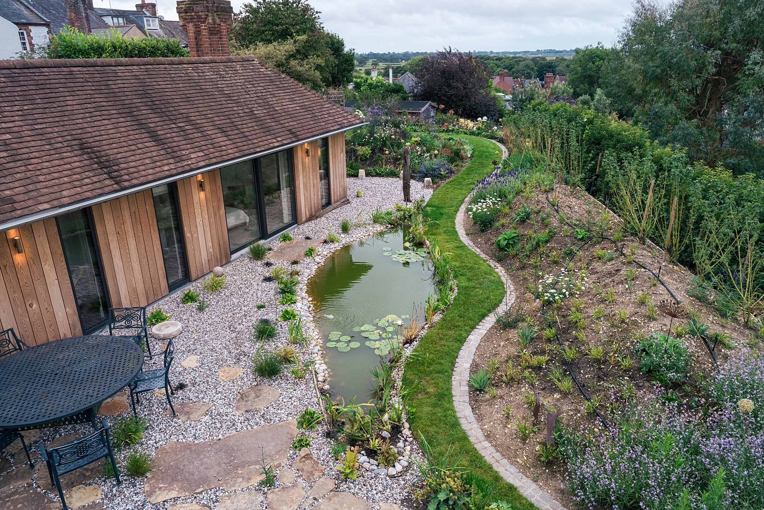 A backyard garden with a pond, surrounded by pebbles and greenery, next to a wooden house with sliding glass doors, and a patio with a black metal table and chairs.