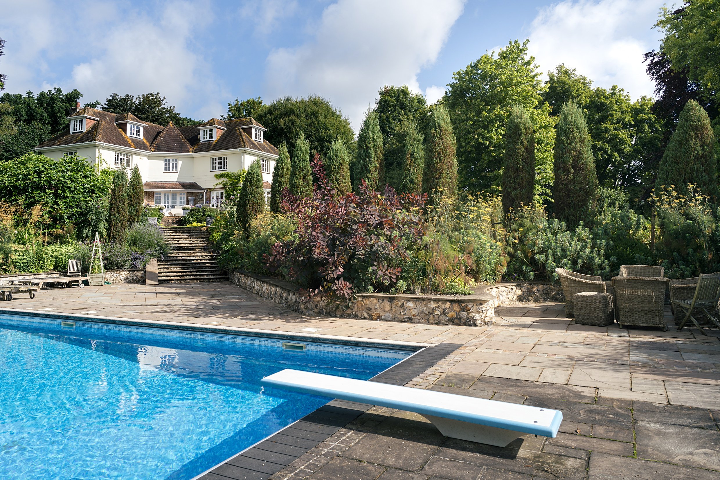 A backyard with a swimming pool, patio furniture, and a garden with a large house in the background surrounded by trees and greenery under a partly cloudy sky.