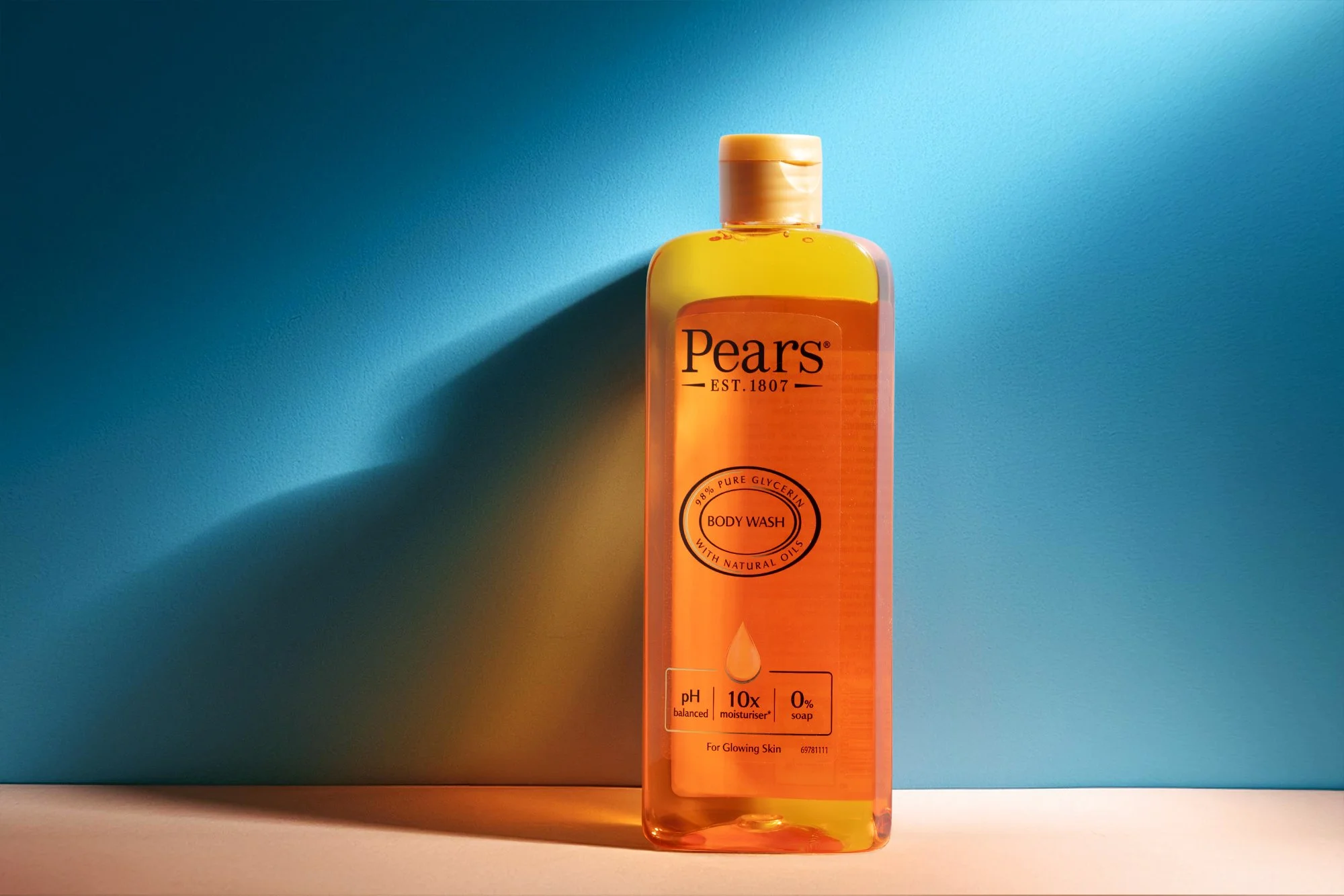 A clear plastic bottle of Pears body wash with yellow and orange liquid, standing on a white surface against a blue background with a shadow cast from the bottle.