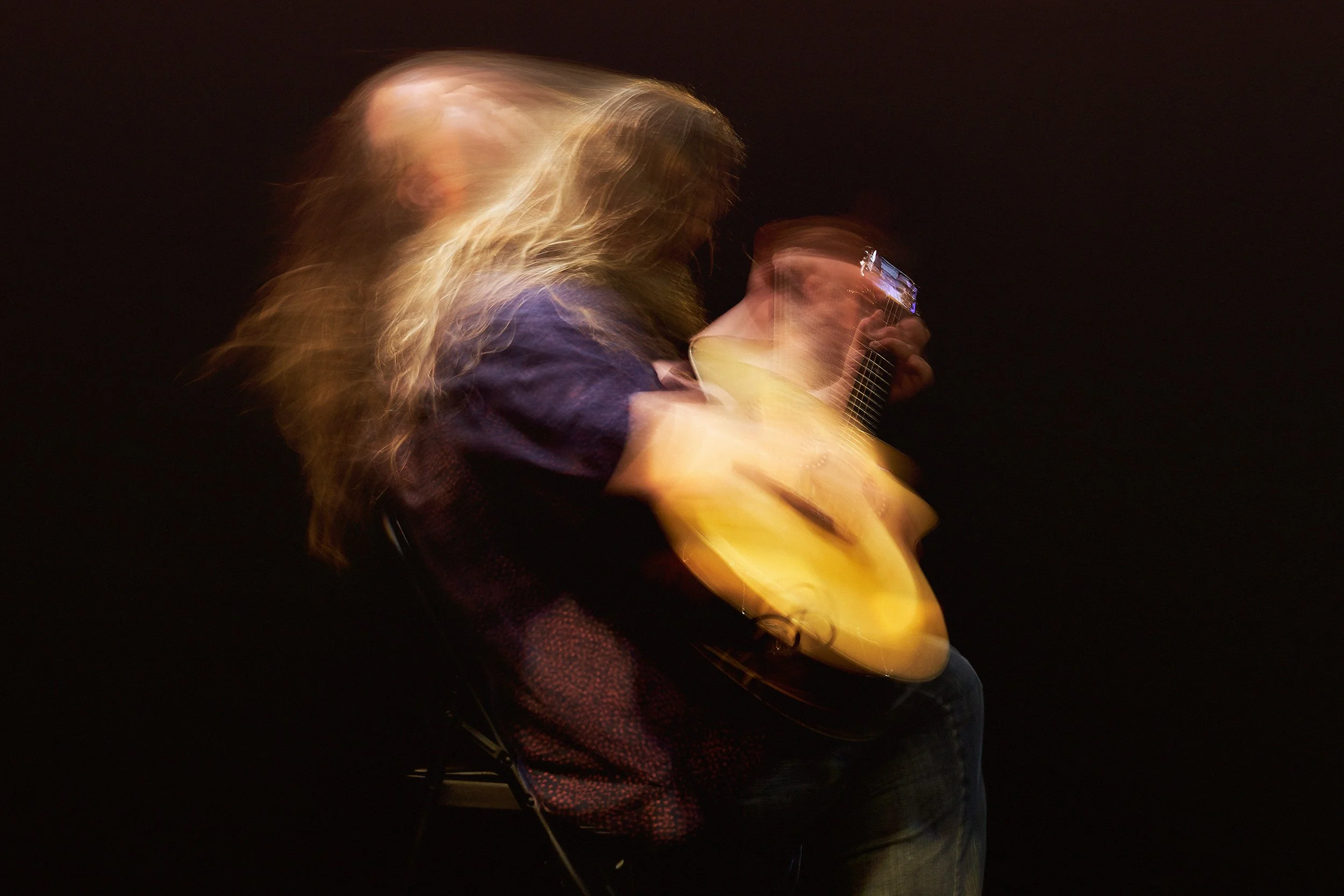 A person with long, blonde hair and a dark shirt is playing an acoustic guitar, with motion blur indicating movement, against a dark background.
