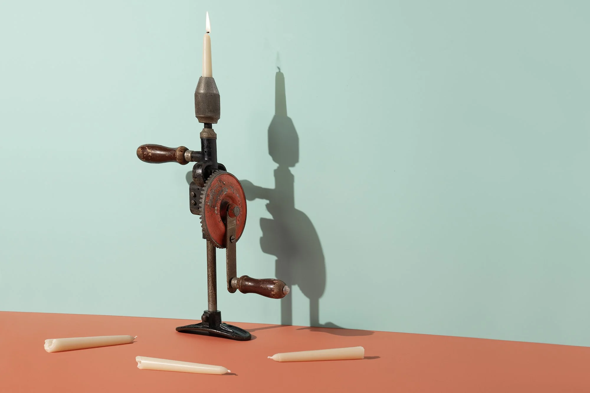 An antique candle-making machine with a red gear, wooden handles, and a metal frame, casting a shadow on a mint green wall. Three white candles are on a coral-colored surface, one standing upright in the machine and two lying flat.