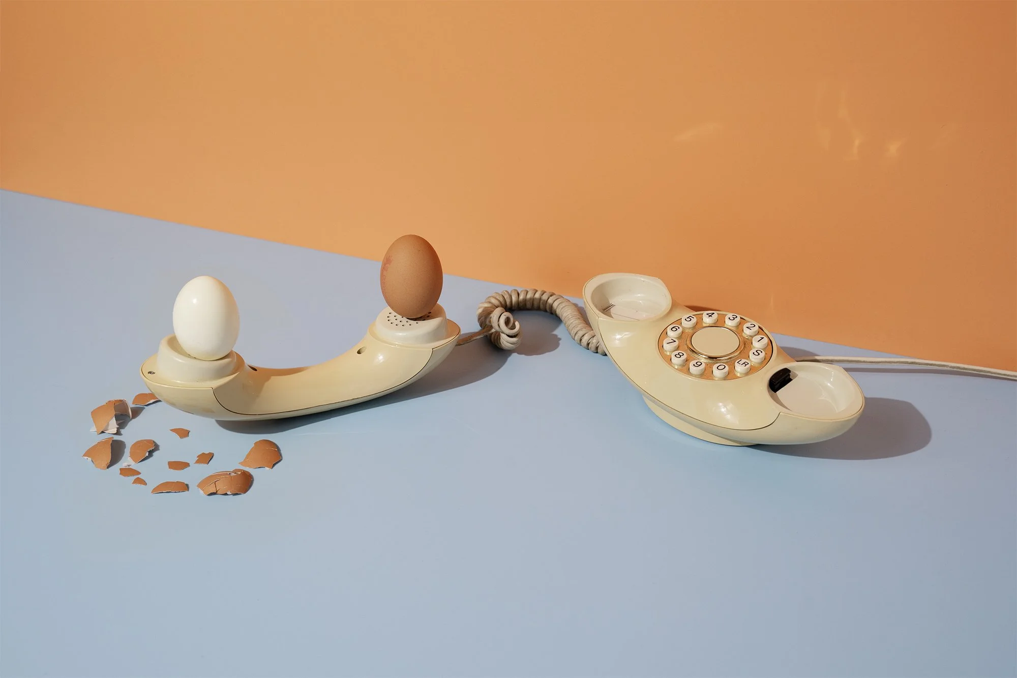 An old beige landline telephone with a rotary dial and a separate receiver, connected by a coiled cord, with two eggs on it and broken eggshells on the surface.
