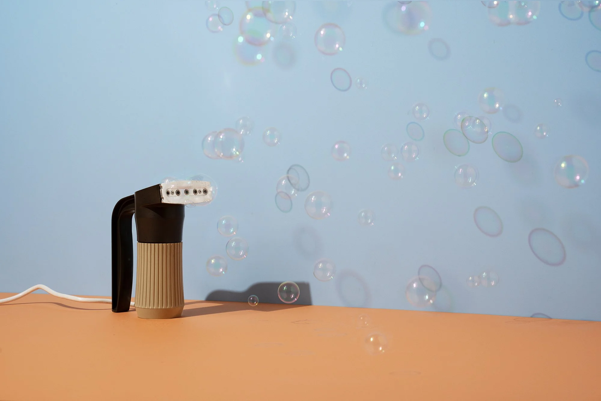 A bubble machine with a beige base and black handle on a peach-colored surface, emitting bubbles against a blue background.