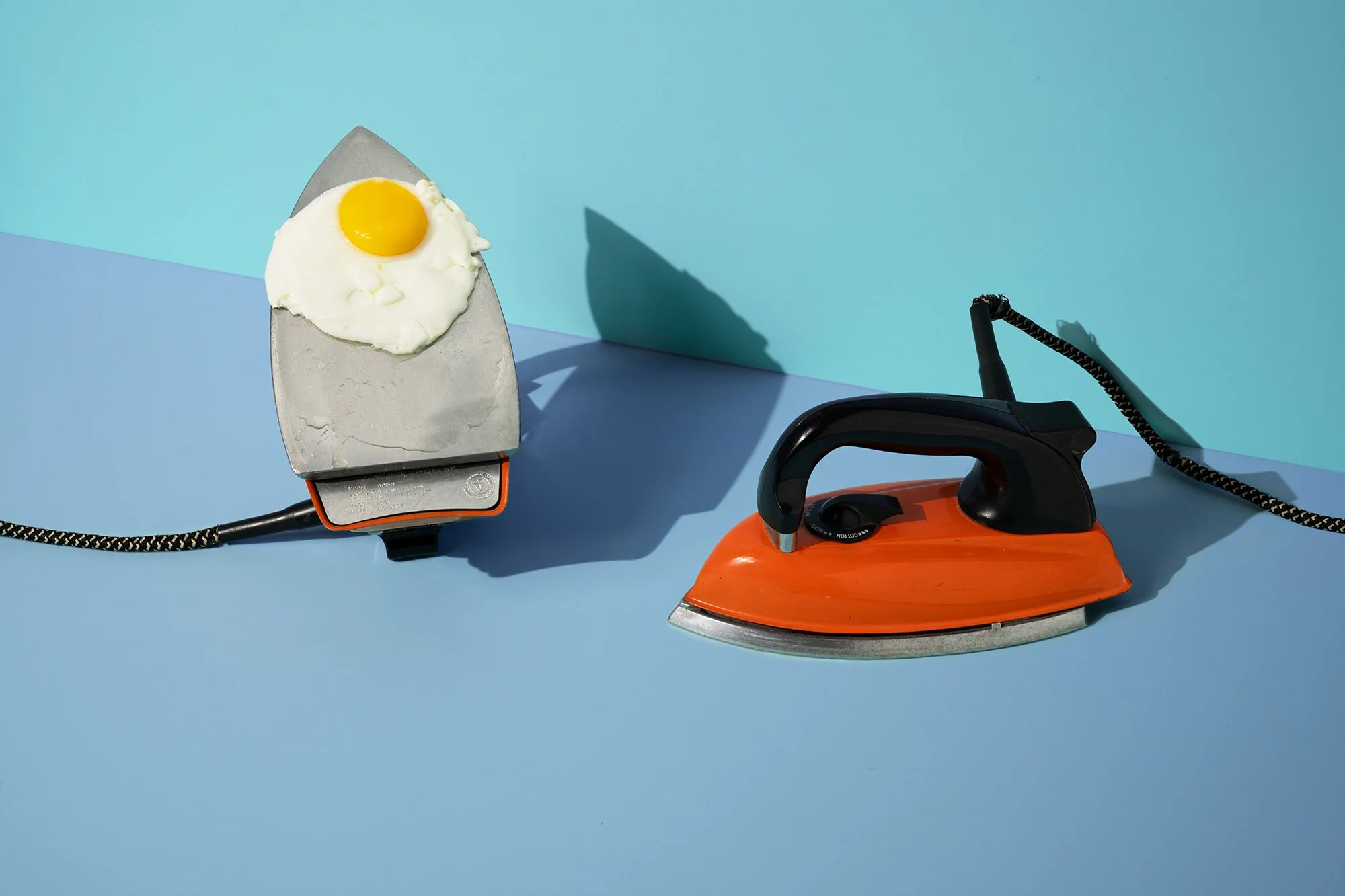 A metallic iron with a fried egg on top, connected to an orange iron by a black and white braided cord, on a blue and cyan background.