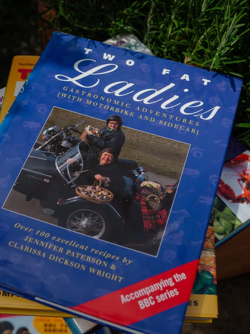 Close-up of the original Two Fat Ladies cookbook cover featuring Jennifer Paterson and Clarissa Dickson Wright beside their motorbike and sidecar.
