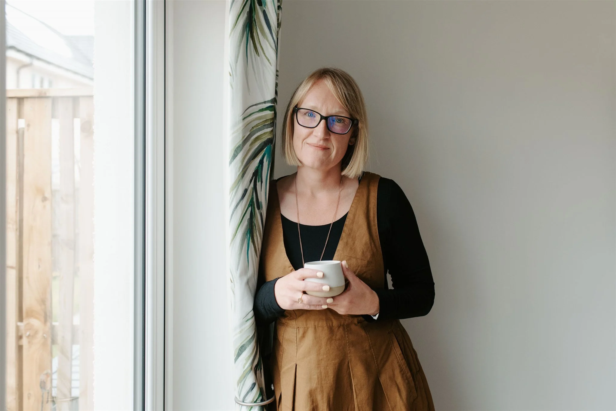 Sarah Philp standing by a window with a warm mug, reflecting on seasonal rhythms and slow living in everyday life.