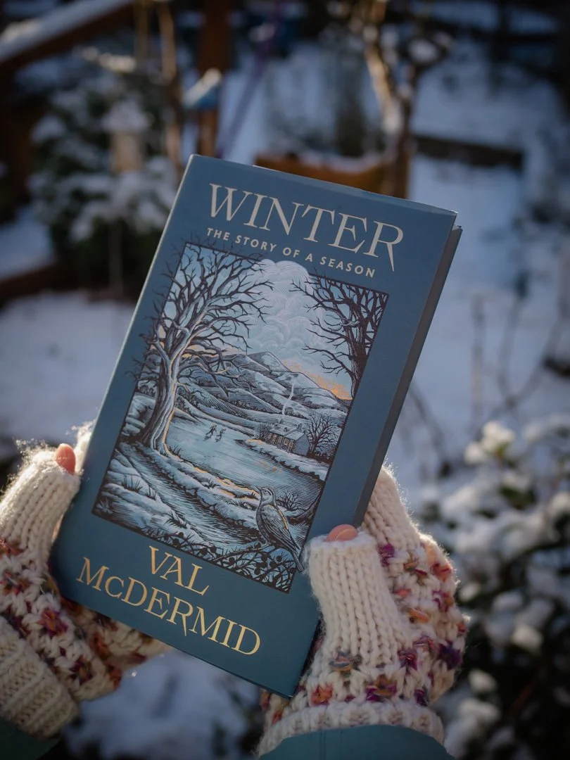 Winter by Val McDermid photographed outdoors in winter, held in gloved hands against a snowy garden backdrop