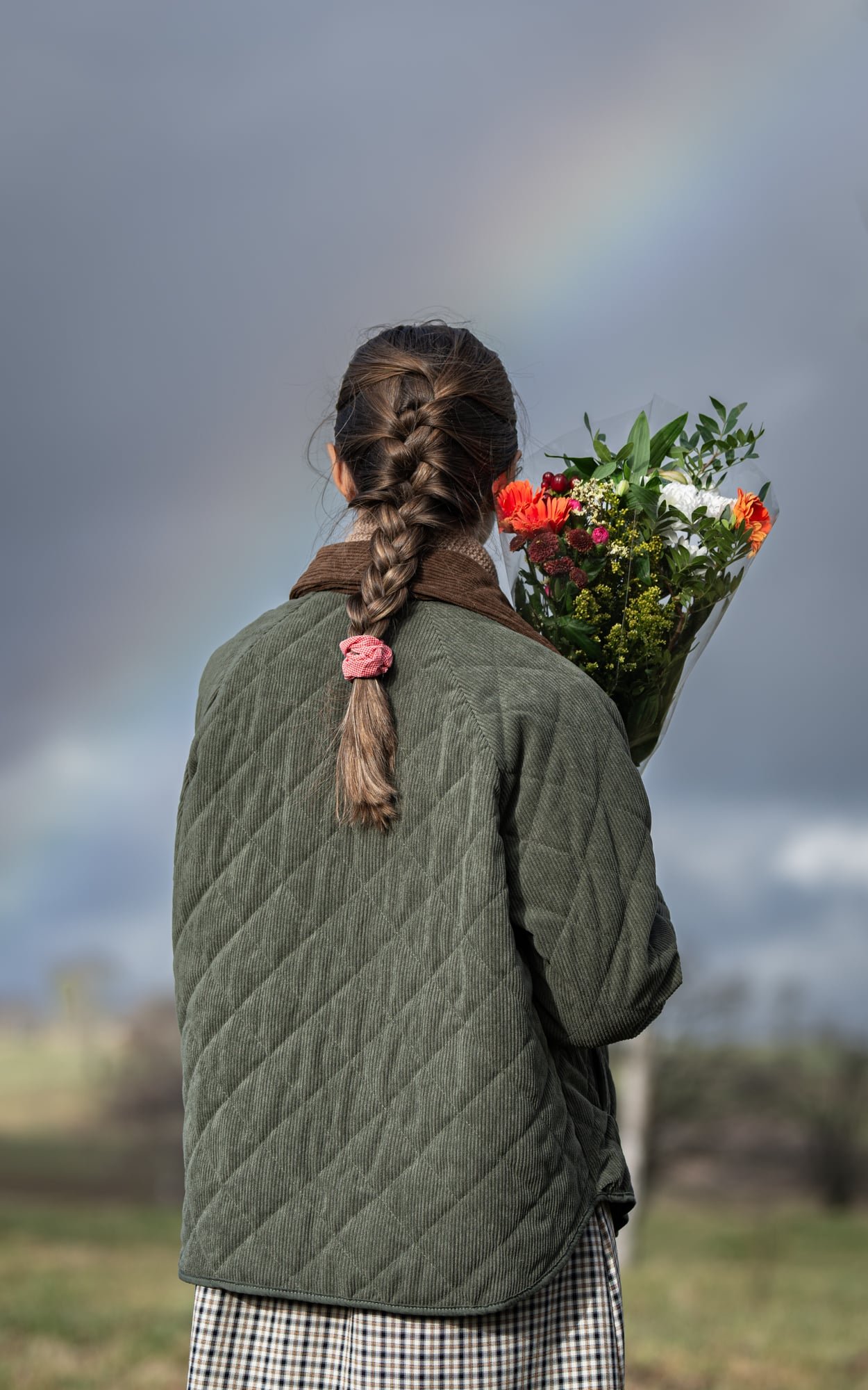 Floriane of The Quiet Days holding a bunch of flowers and looking towards a rainbow in a dark countryside sky.