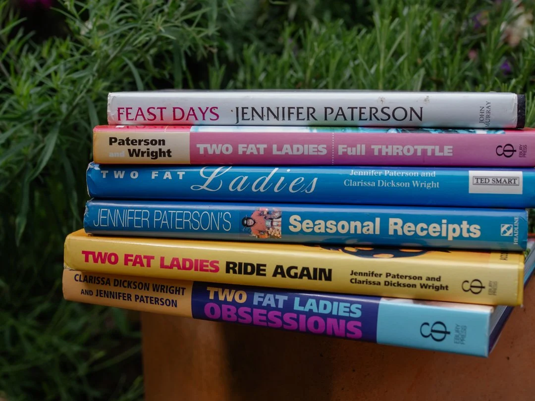 A stack of Two Fat Ladies cookbooks photographed from a different angle, showcasing classic British cookery books.