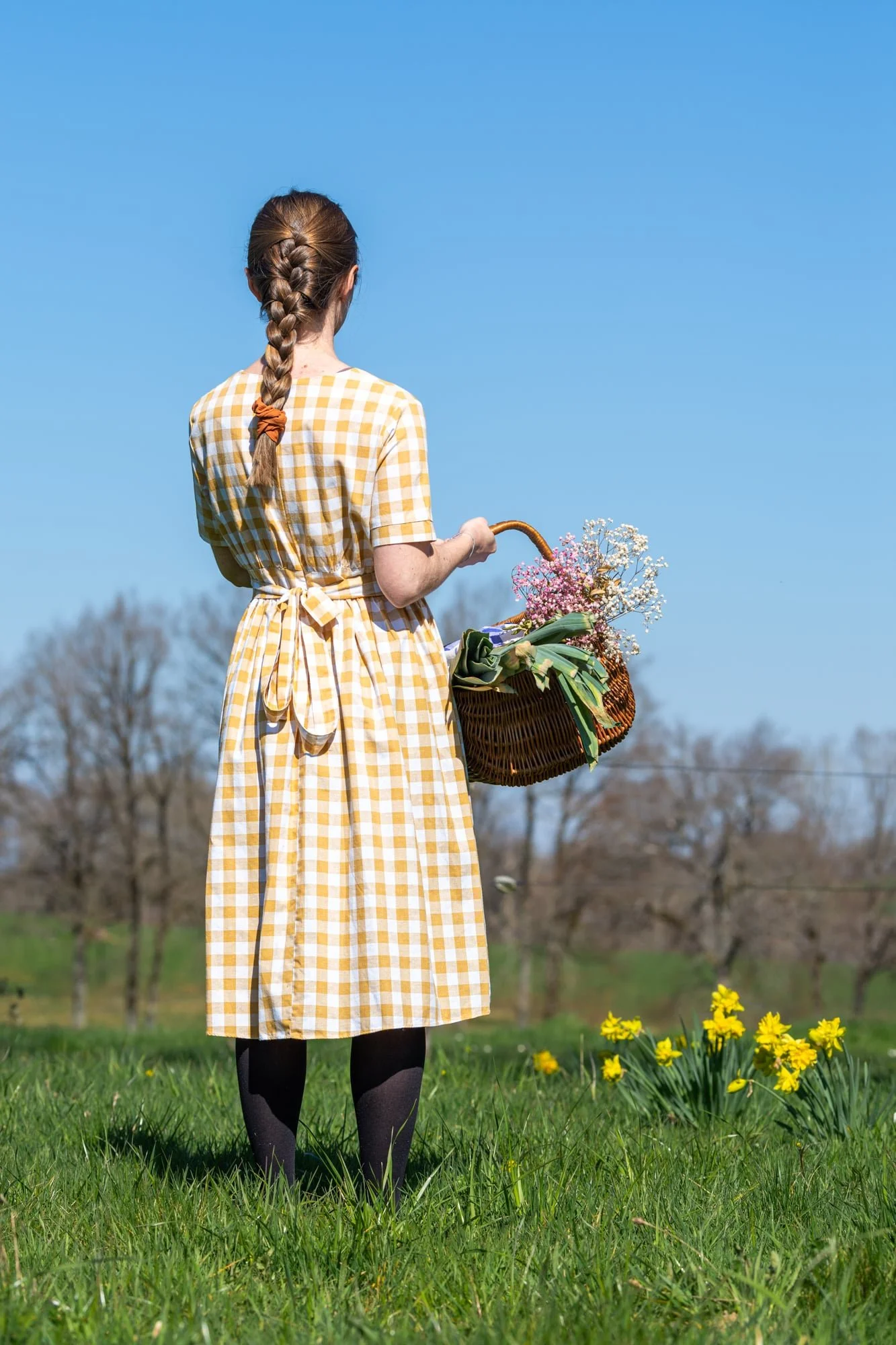 Floriane of The Quiet Days wearing a yellow gingham dress and holding a basket of seasonal wildflowers, herbs and vegetables outdoors in summer.