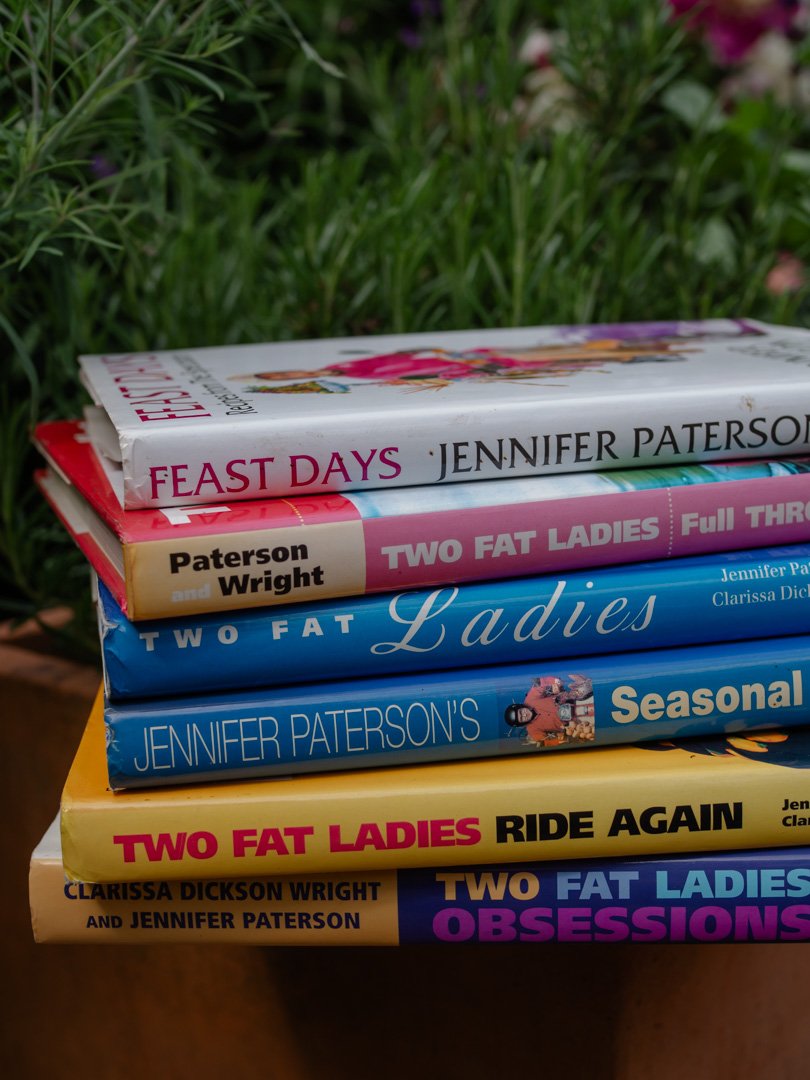 A collection of Two Fat Ladies cookbooks by Jennifer Paterson and Clarissa Dickson Wright stacked on a table.