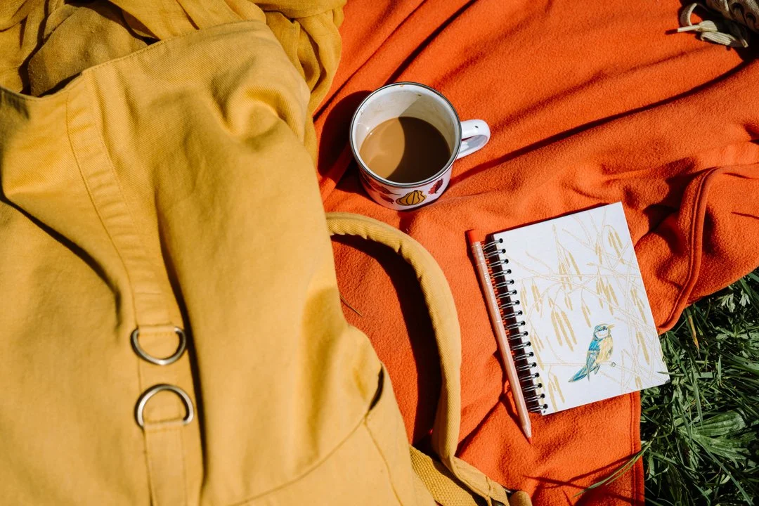 A sketchbook, rucksack and mug of hot tea on a blanket outdoors, representing slow, simple and seasonal living.