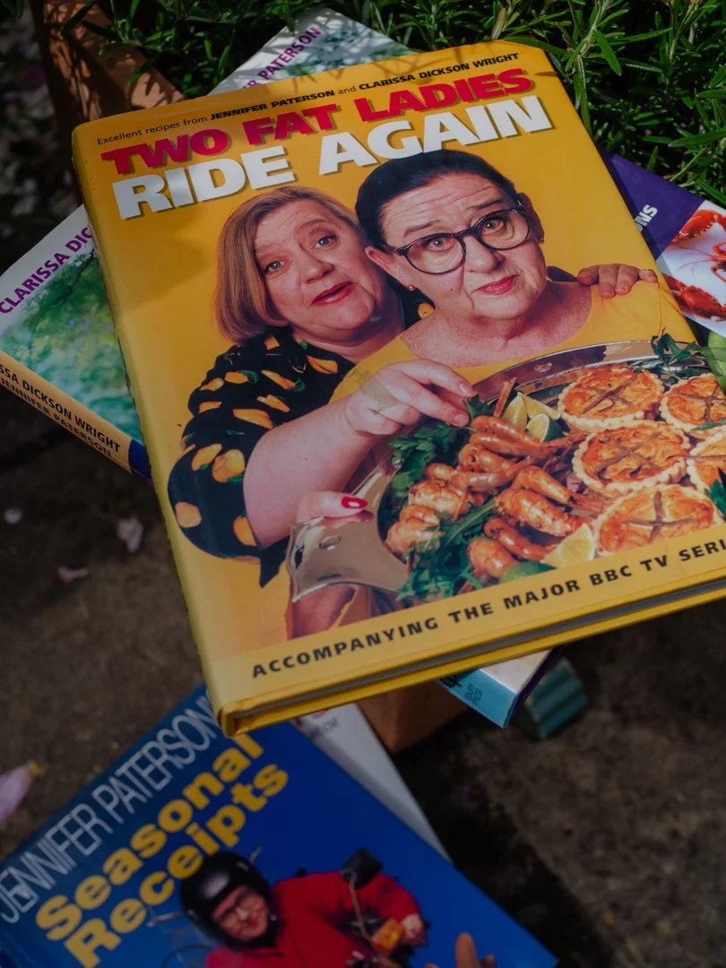 The Two Fat Ladies Ride Again cookbook displayed on top of a collection of cookbooks by Jennifer Paterson and Clarissa Dickson Wright.
