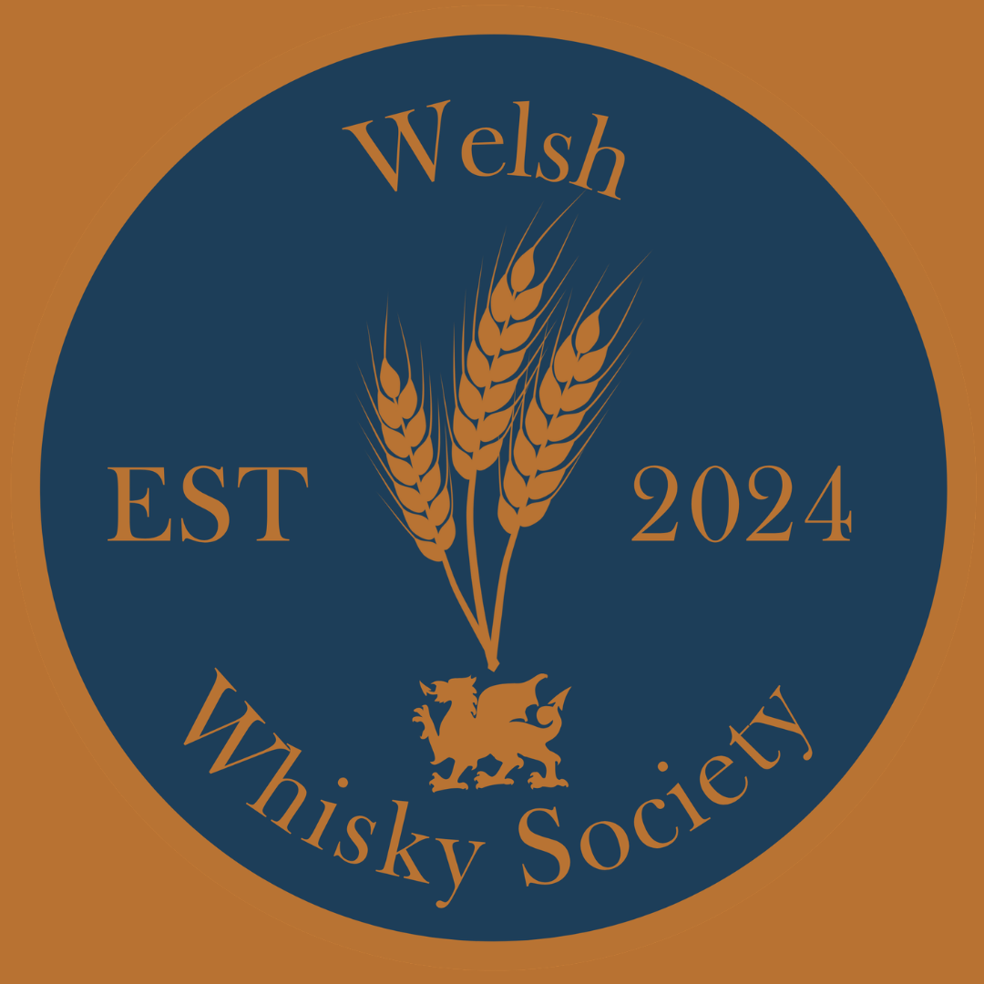 A circular emblem with a blue background and brown border. It features three wheat stalks at the center. Above, it says "Welsh" in brown text. To the left, "EST" and to the right, "2024" in brown. Below, it reads "Whisky Society" in brown text, with a small stylized lion emblem beneath the wheat stalks.
