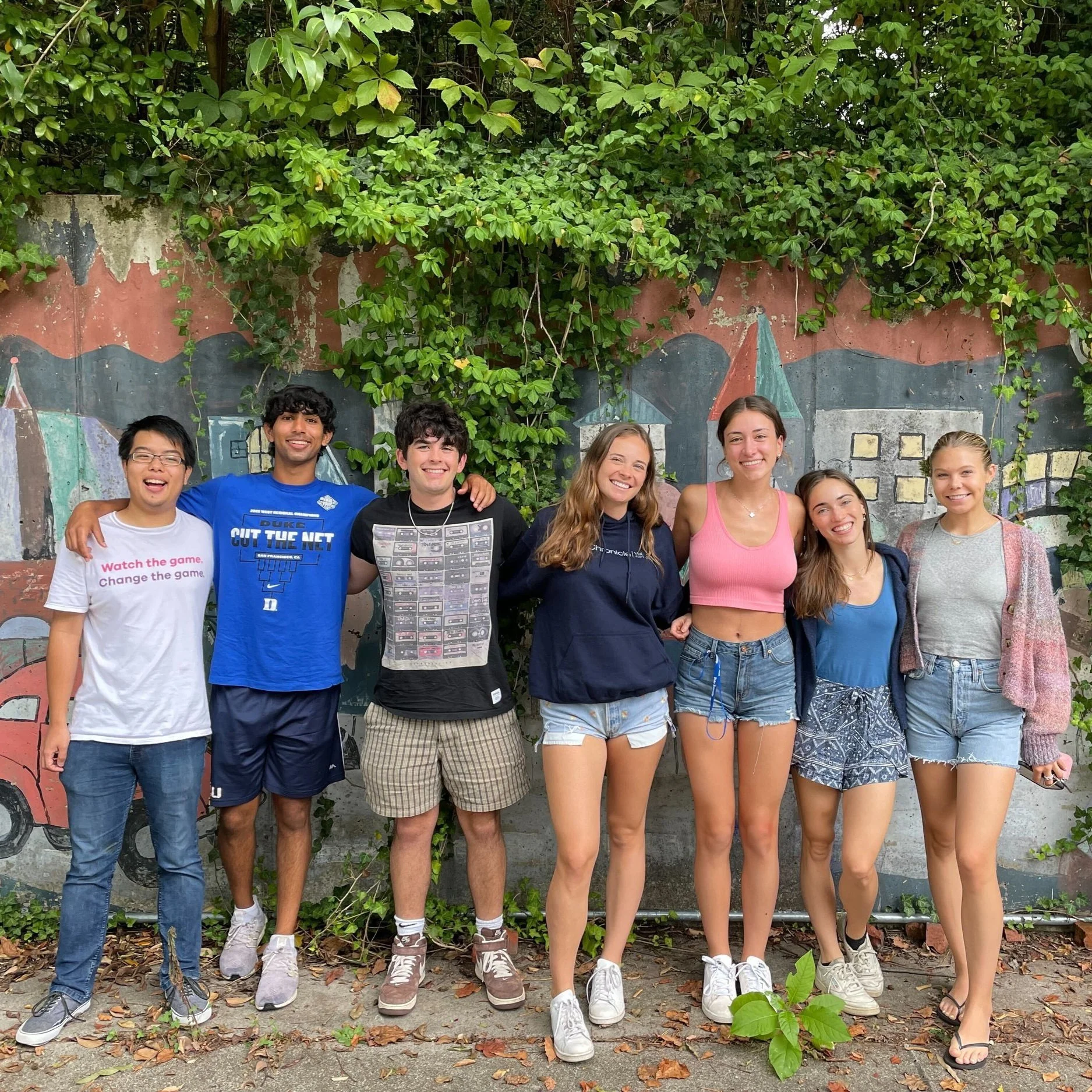 The students of the Ad Team lined up in front of a city mural and green vines and leaves.