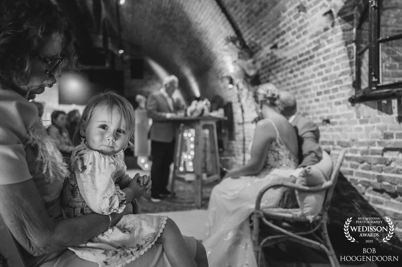 Foto van een groep mensen, inclusief een jonge meisje die op de schoot zit van een vrouw, in een rustieke ruimte met bakstenen muren, mogelijk bij een bruiloft of evenement.