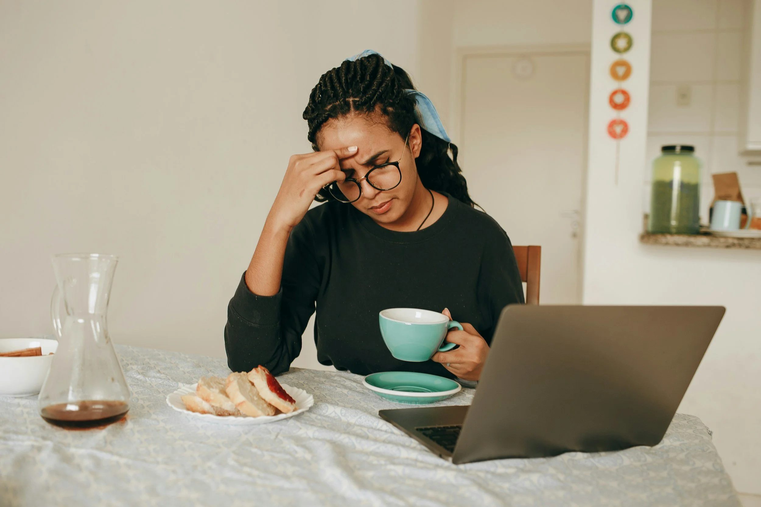 Woman sitting at a table with a laptop and coffee, holding her forehead while looking overwhelmed.