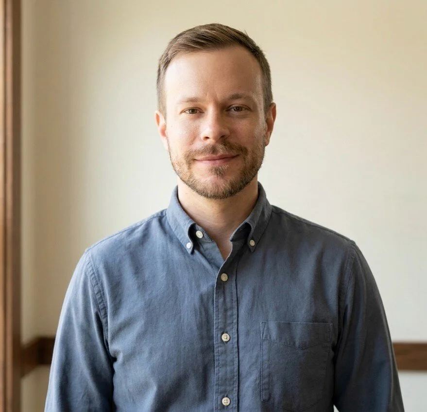 A young man with short brown hair, a beard, wearing a gray button-up shirt, standing indoors with a neutral expression.