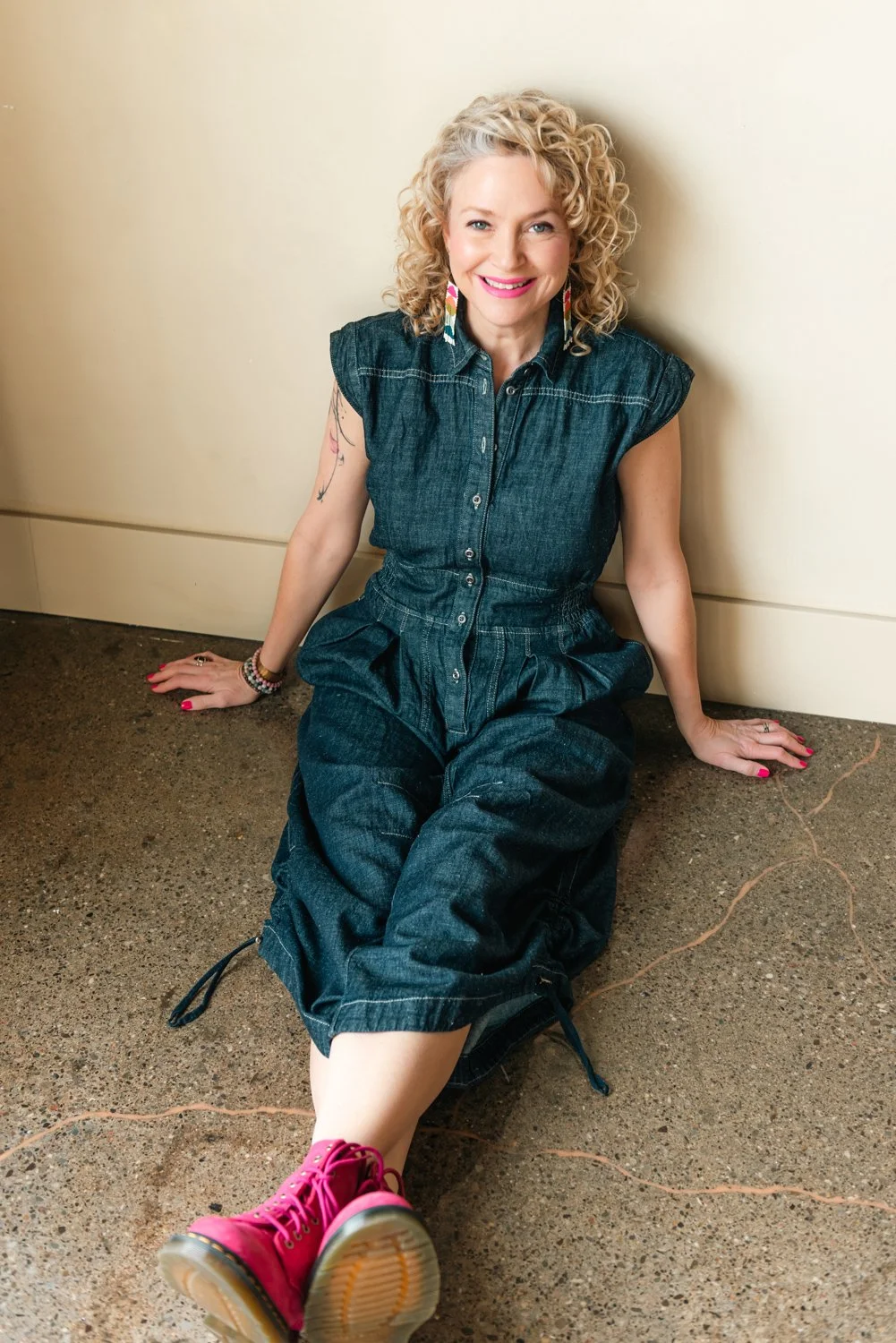Laura Beauparlant with curly blonde hair, dressed in a denim jumpsuit, sitting on the floor and smiling, wearing pink boots and colorful earrings, leaning against a wall.