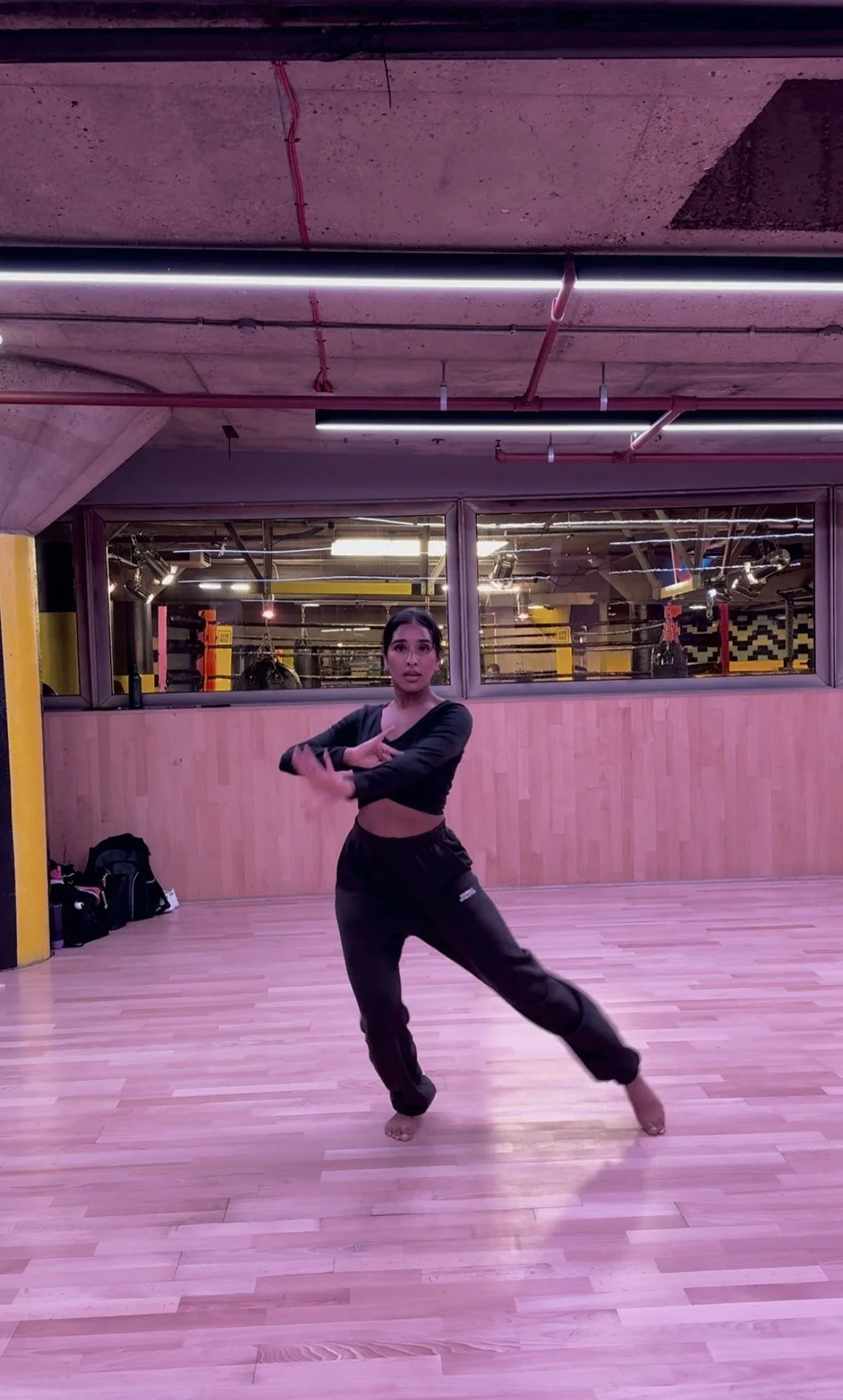 A woman in black workout clothes dancing or performing a pose in a dance studio with wooden floors and a large mirror behind her.