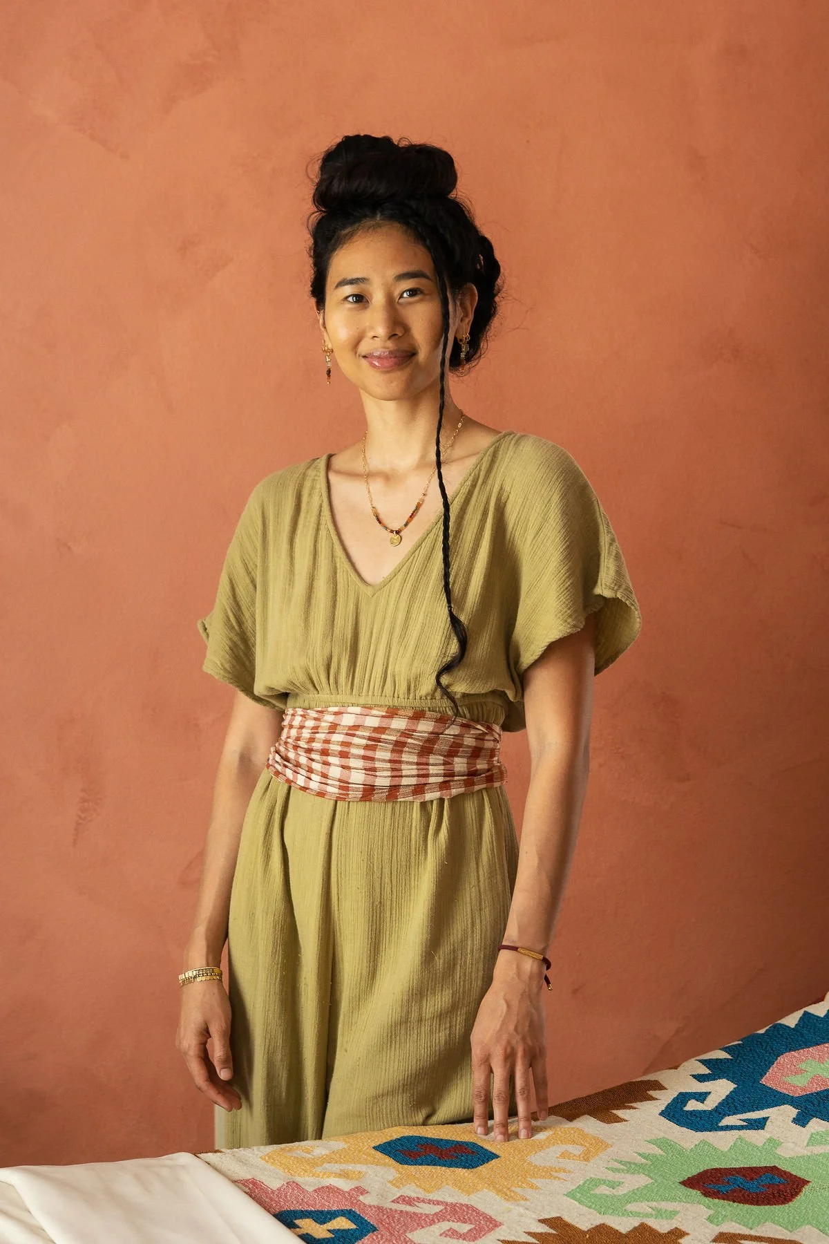 A woman with dark hair styled in a top knot and a single long braid, wearing a yellow dress with a red and white checkered sash around her waist, standing against a peach-colored wall. She has jewelry including earrings, necklaces, and bracelets, and is smiling gently.