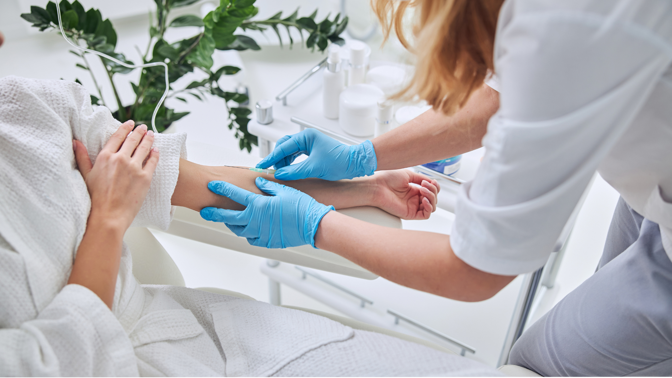 A nurse in blue gloves administering an injection into a patient's arm in a bright medical room with white shelves and green plants.