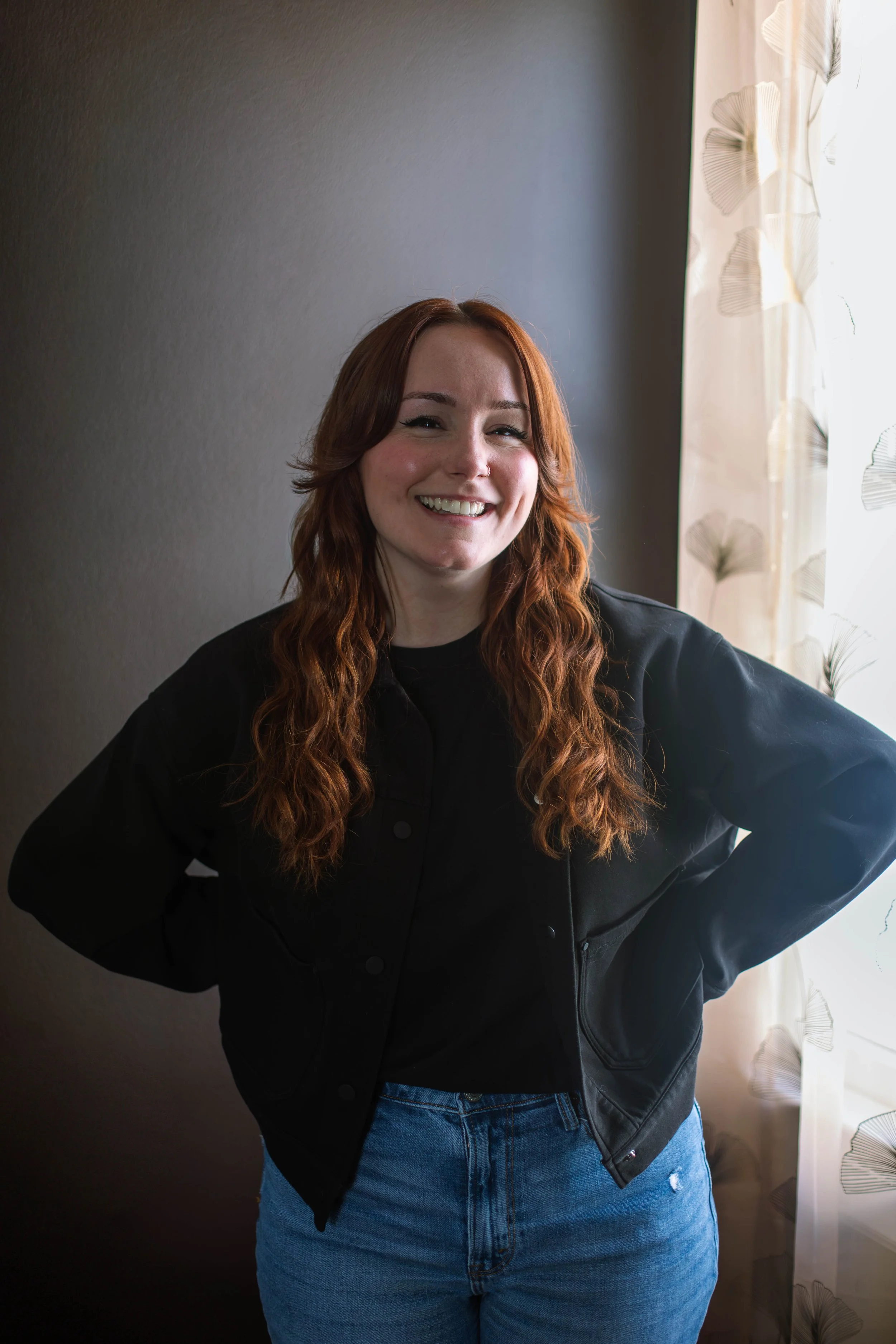 A smiling woman with wavy red hair and a black jacket posing confidently against a dark wall in soft, natural window light.