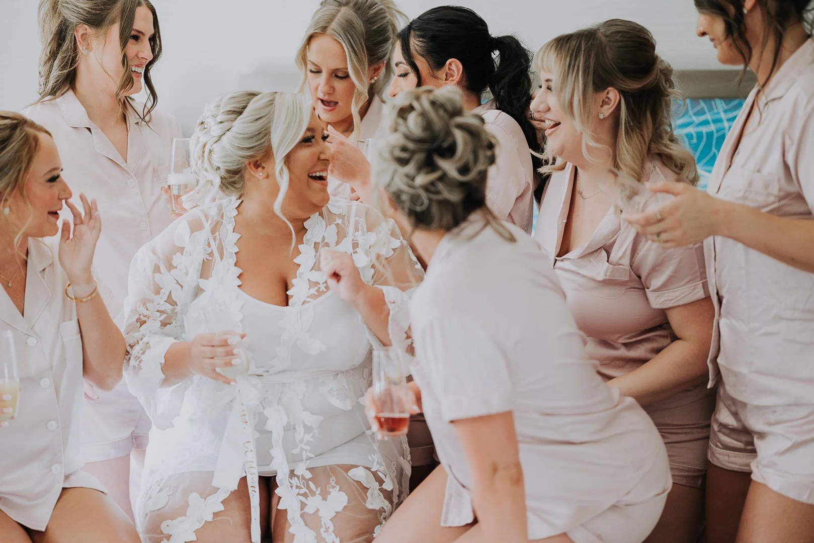 Group of women in matching light pink pajamas celebrating a bride in a white dress, with some women holding glasses of champagne, smiling and laughing in a joyful moment.